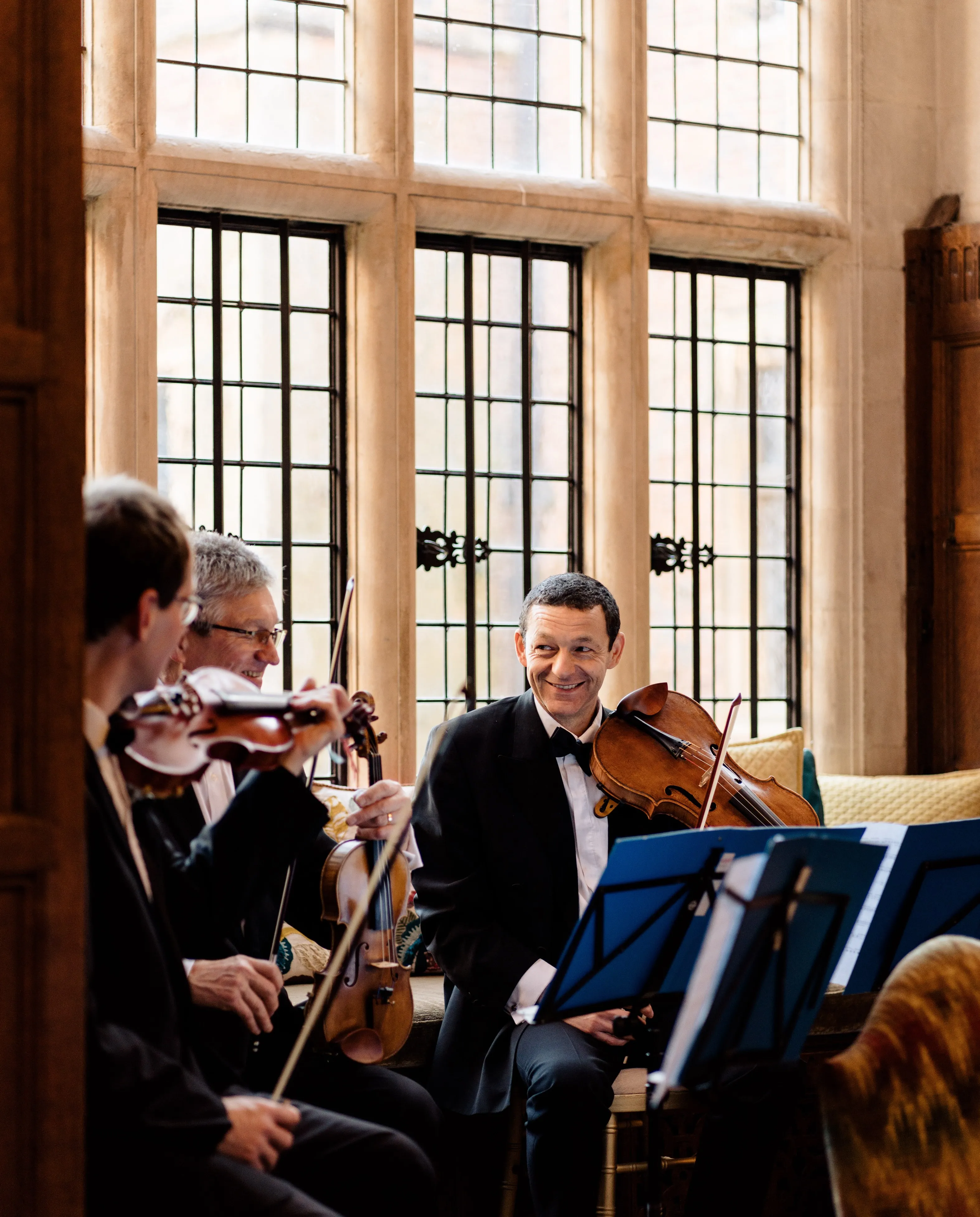 Three violinists happily playing wedding songs in the magnificent high gallery, surrounded by stunning floor-to-ceiling windows. The musicians' joyful expressions are visible as they serenade the wedding guests with their melodious tunes. The grand gallery's opulent architecture and the abundant natural light streaming in through the impressive windows add to the enchanting ambiance of the wedding ceremony, creating a memorable and elegant setting