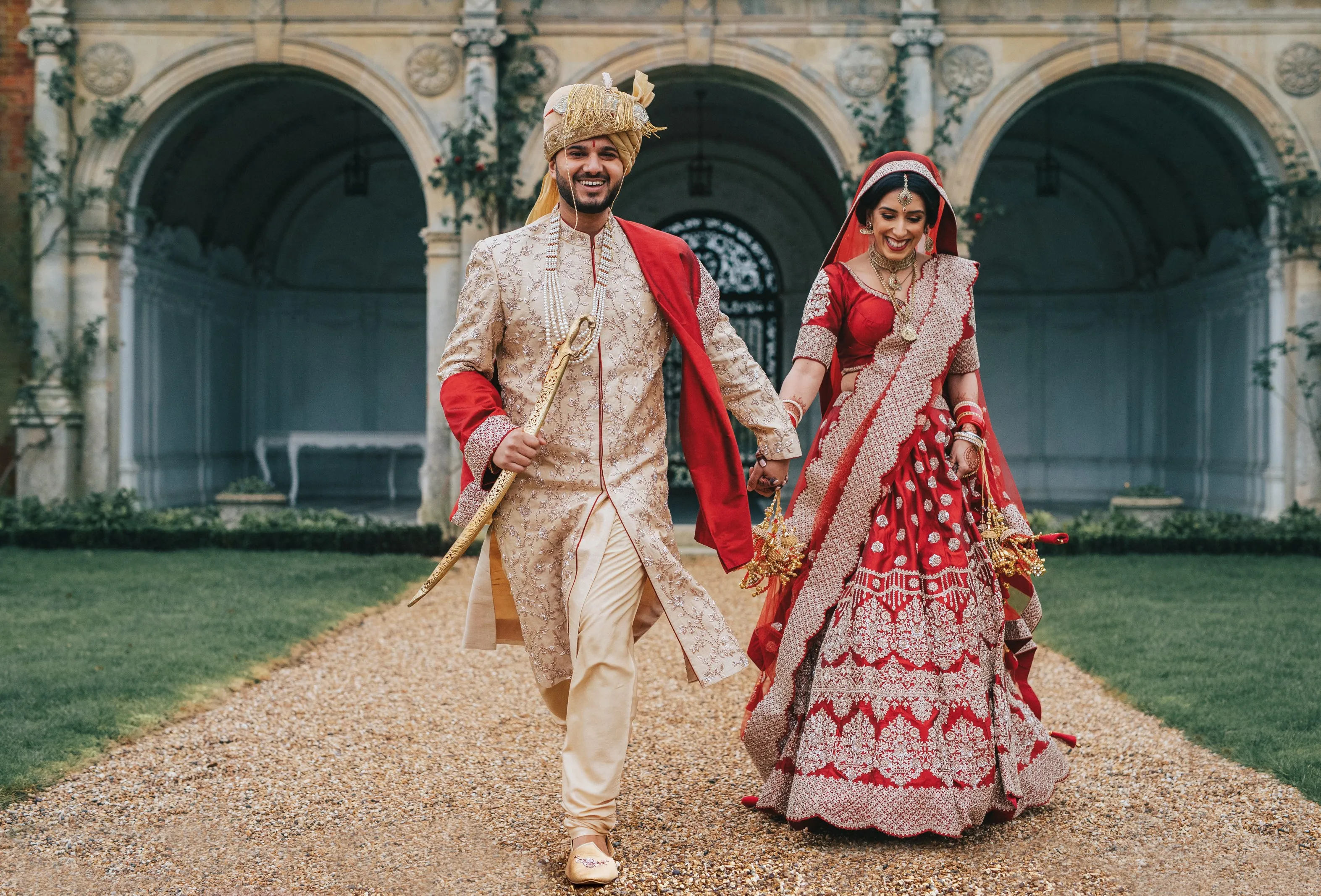A happy Asian bride and groom, dressed in vibrant red traditional outfits, walking hand in hand in the beautiful gardens of North Mymms Park, with the Loggia in the background. The couple's radiant smiles reflect their joy as they embark on their new journey together. The picturesque gardens, lush with greenery and colorful blooms, complement the couple's attire, creating a stunning and joyful scene for their special day