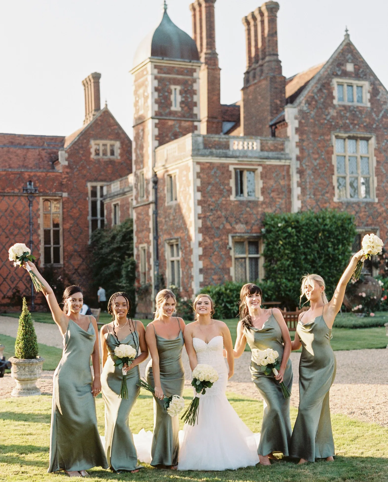 he bride and her bridesmaids standing together in the beautiful gardens of North Mymms Park, raising their hands and flowers in the air with happiness. The lush greenery and colorful blooms provide a picturesque setting for this joyful moment. The majestic mansion house serves as a stunning backdrop, adding to the charm and elegance of this cheerful scene. The bride and her bridesmaids' laughter and excitement create a memorable and heartwarming atmosphere on this special day