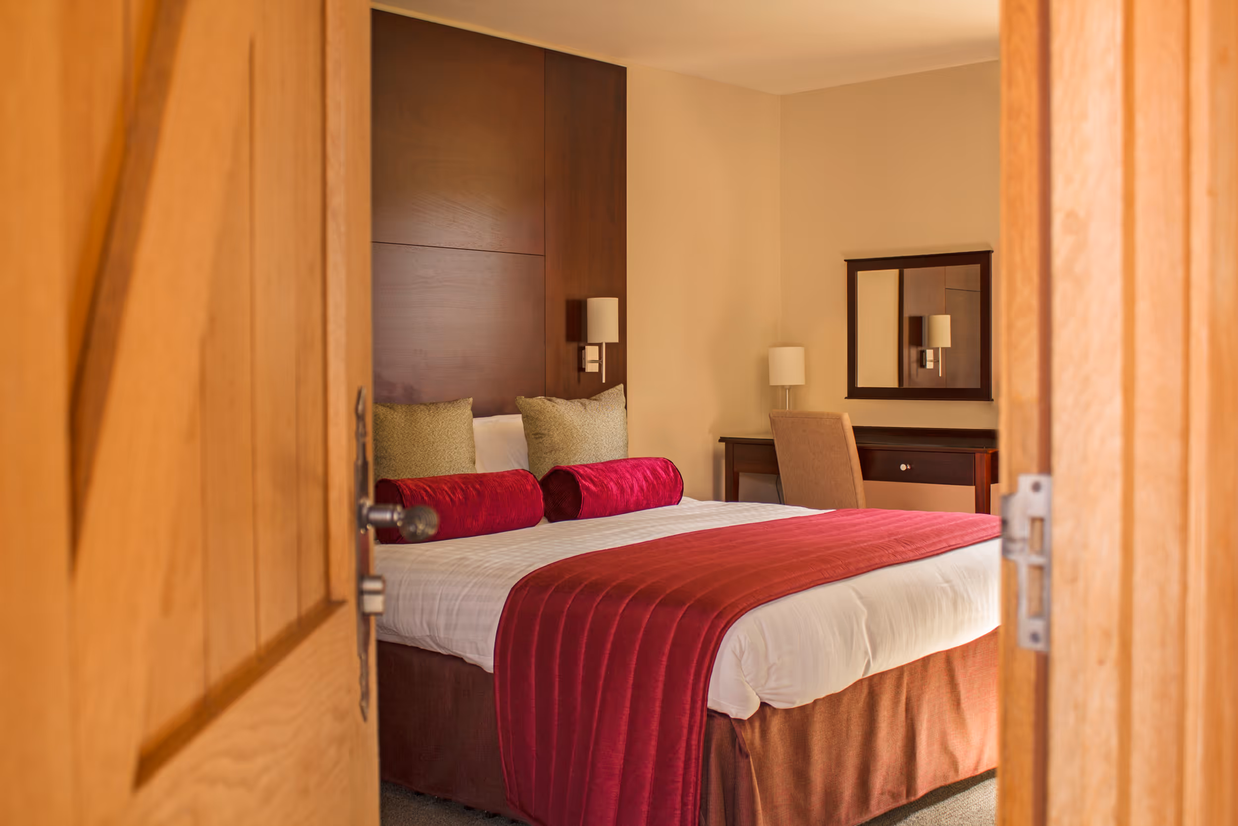 A modern guest bedroom in the stables at North Mymms Park. The room is equipped with a comfortable double bed with cushions, a sleek TV, a convenient dressing table, and coffee facilities. Large mirrors adorn the room, and an en-suite bathroom completes the inviting setup, providing a perfect space for guests to relax and unwind.