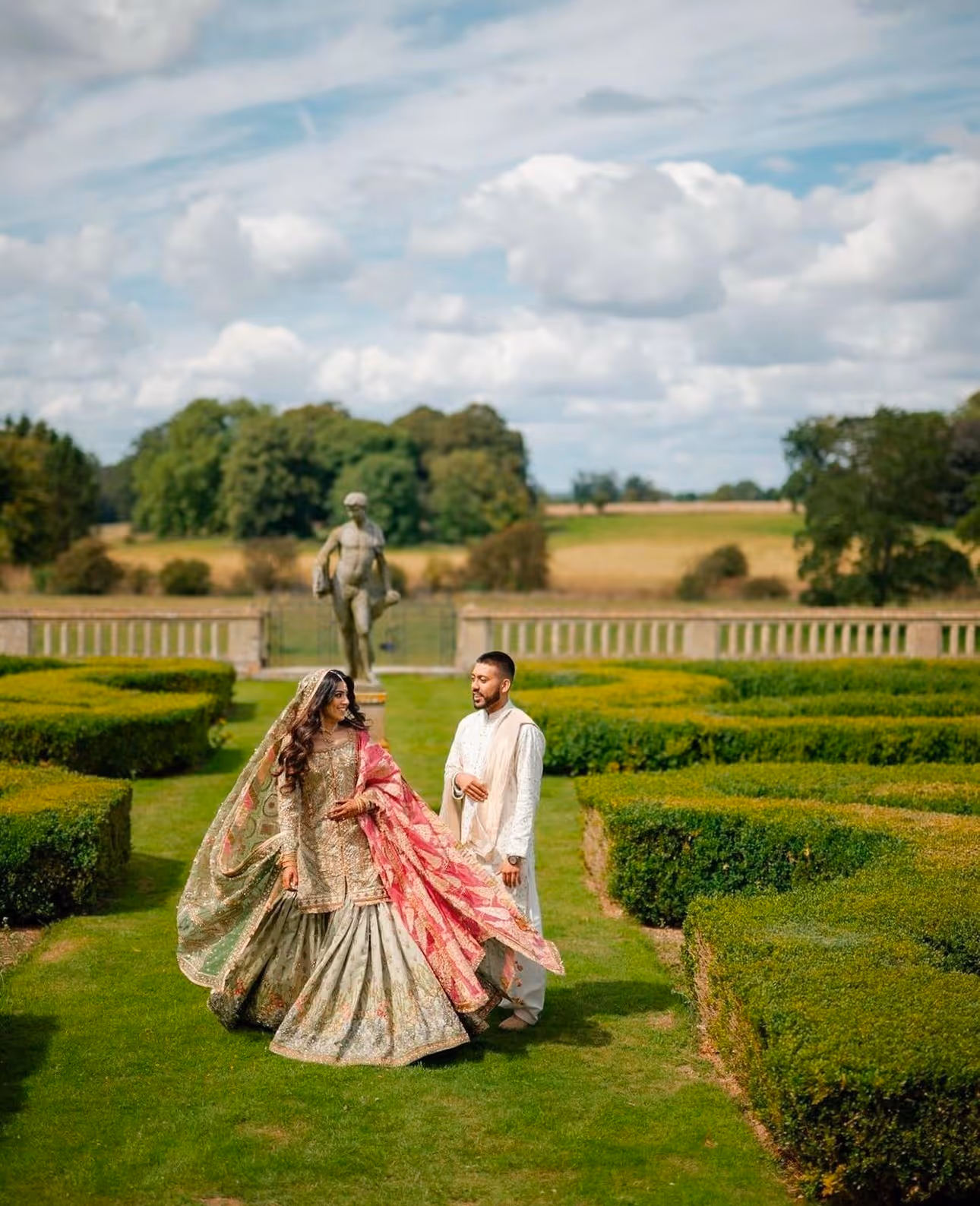 Stunning Asian bride and groom posing in the beautiful gardens of North Mymms Park, with a historic statue gracefully standing in the background, adding a timeless touch to their romantic portrait