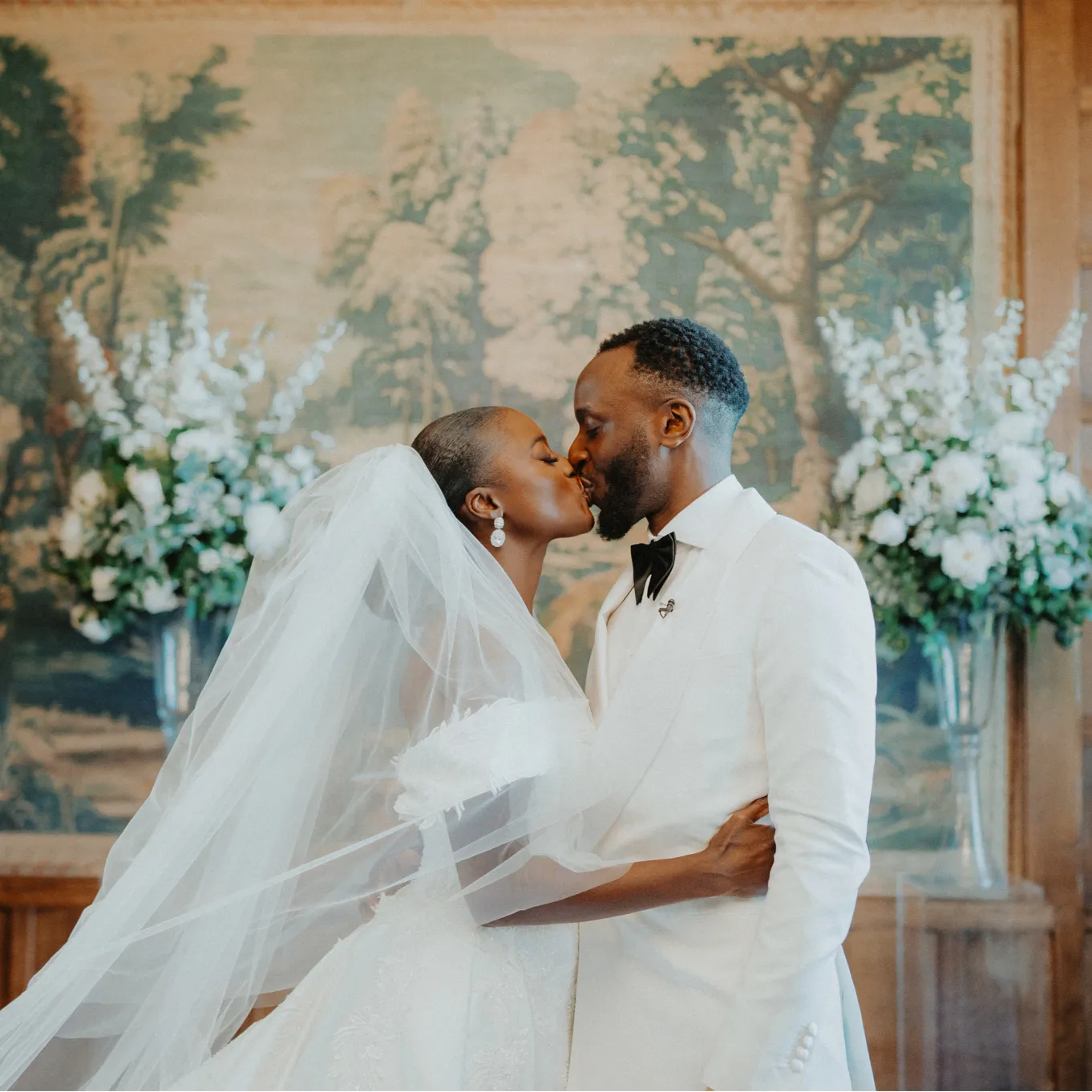 An African couple sharing their first kiss after a heartwarming civil ceremony at North Mymms Park. They stand in front of a radiant tapestry, with vibrant flowers arranged beautifully on both sides of them. The moment is filled with love and joy, celebrating their union in an elegant and picturesque setting