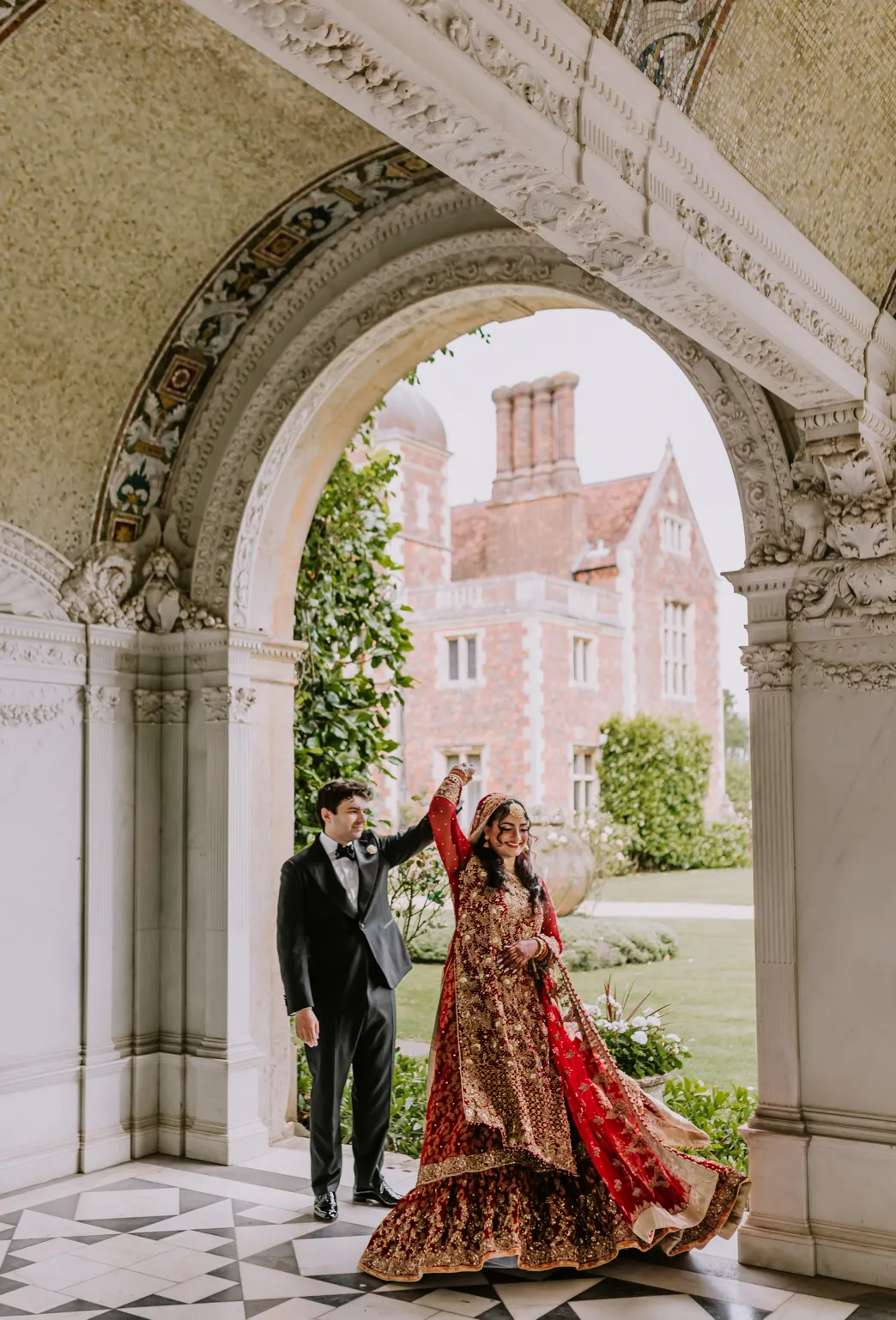 A joyful Asian bride and groom dance together in a stunning loggia, their movements filled with happiness and love. Behind them, the majestic North Mymms Park provides a breathtaking backdrop, enhancing the beauty and romance of the moment