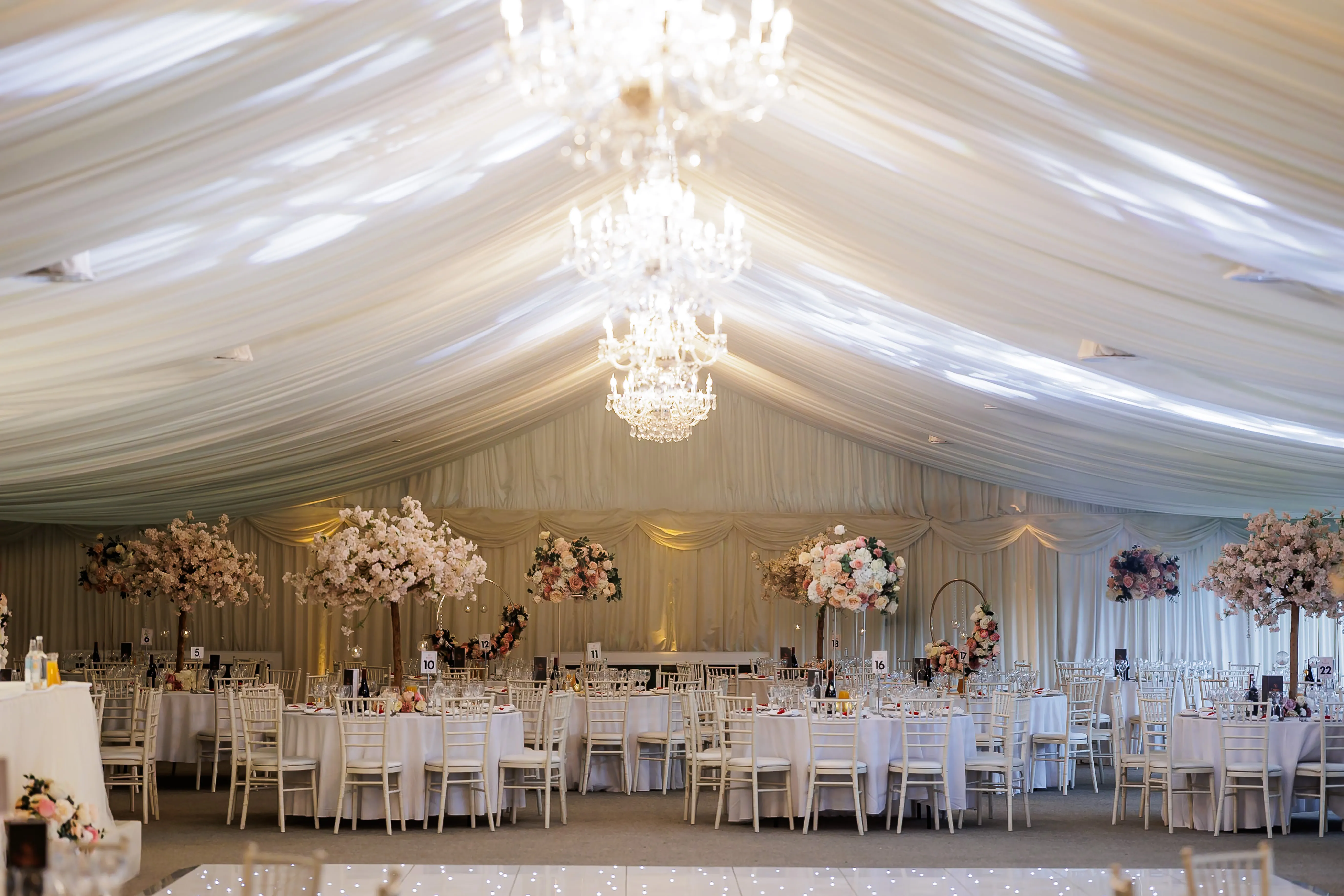 Elegant wedding reception setup inside a beautifully draped marquee with crystal chandeliers. The room features round tables covered in white linens, adorned with tall floral centerpieces of pink and white blossoms. White Chiavari chairs surround the tables, and a twinkling LED dance floor is visible in the foreground, creating a romantic and sophisticated atmosphere in North Mymms Park's Pavilion.
