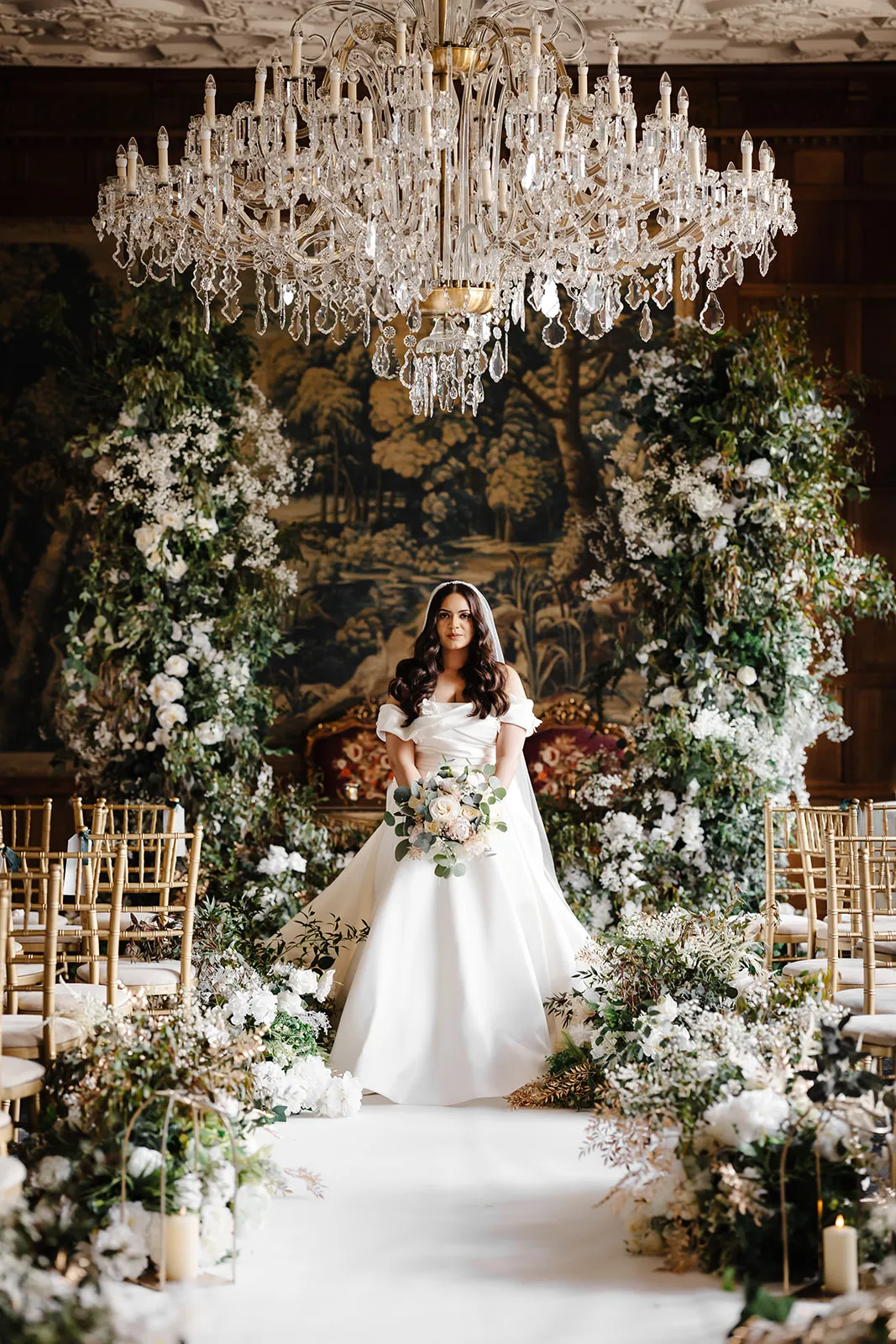 Elegant bride standing among lush florals beneath a large, ornate chandelier in the magnificent Long Gallery at North Mymms Park.