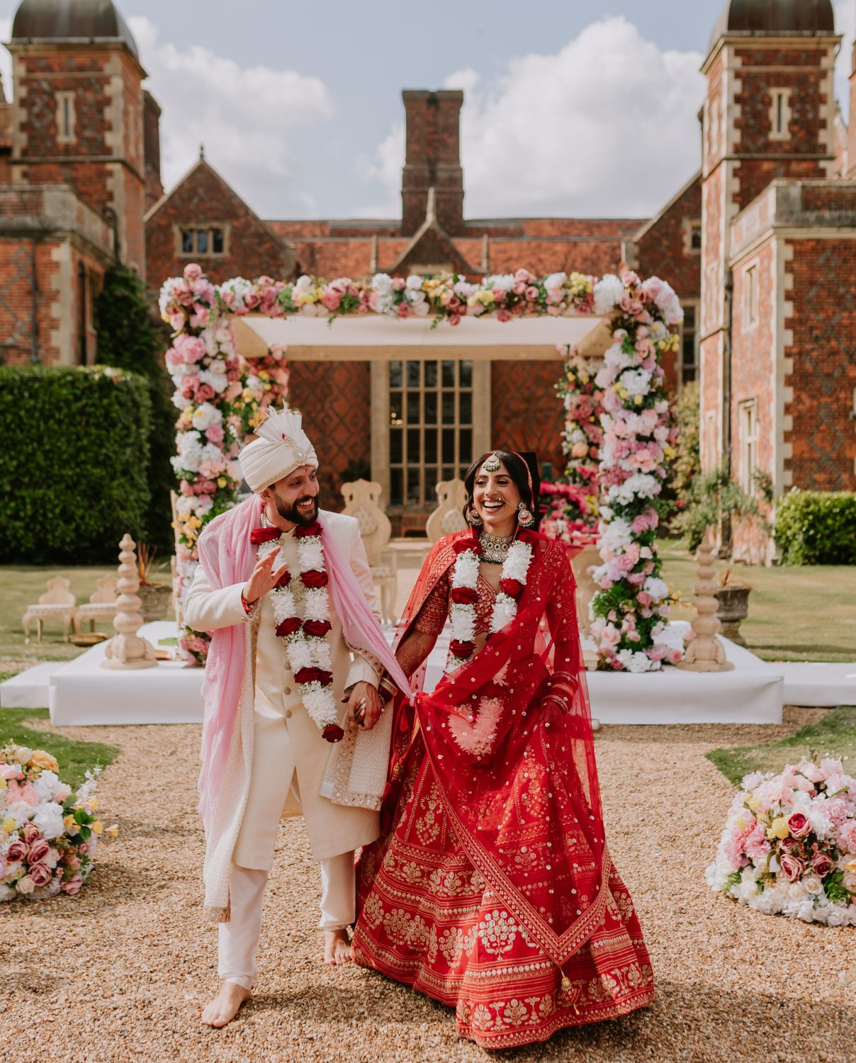 A smiling bride and groom in traditional South Asian wedding attire walk hand-in-hand along a gravel path after their Mandap ceremony at North Mymms Park.