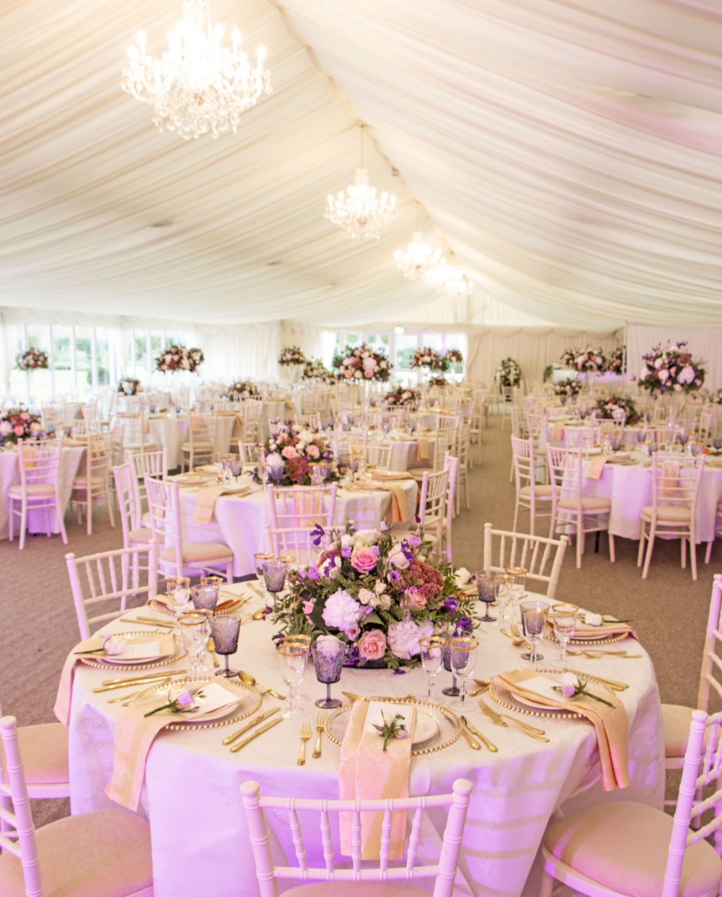 The Pavilion at North Mymms Park dressed for a wedding reception. Round tables are styled with gold chargers, purple glassware, peach napkins, and lush pink and purple floral centrepieces. White Chiavari chairs surround the tables beneath a white draped marquee ceiling with elegant crystal chandeliers.