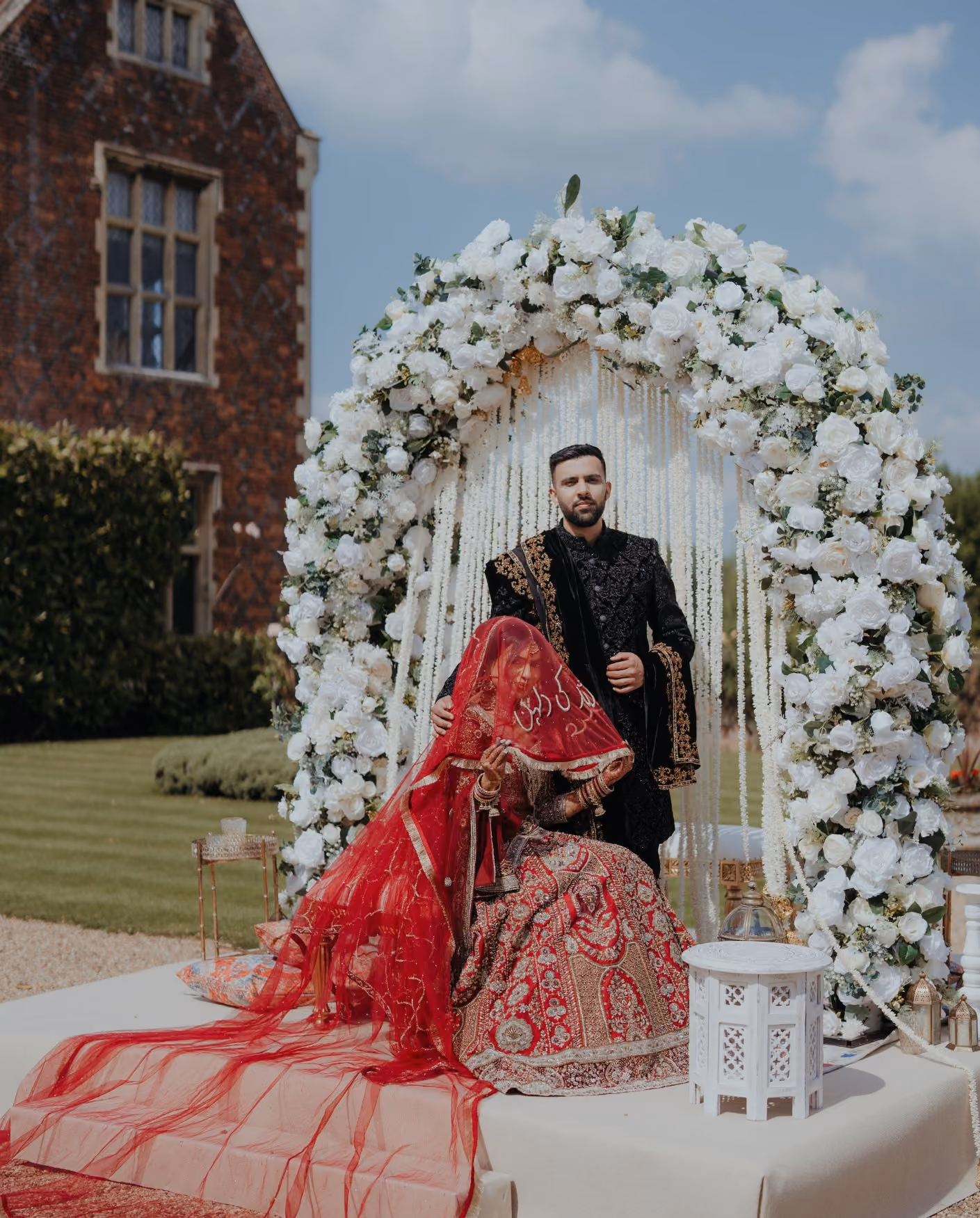 An outdoor Nikah setup featuring a bride in a red embroidered lehenga and veil seated beside her groom in a black sherwani. They pose on a white stage beneath a large arch of white roses with a white beaded backdrop, set against the historic manor house at North Mymms Park.