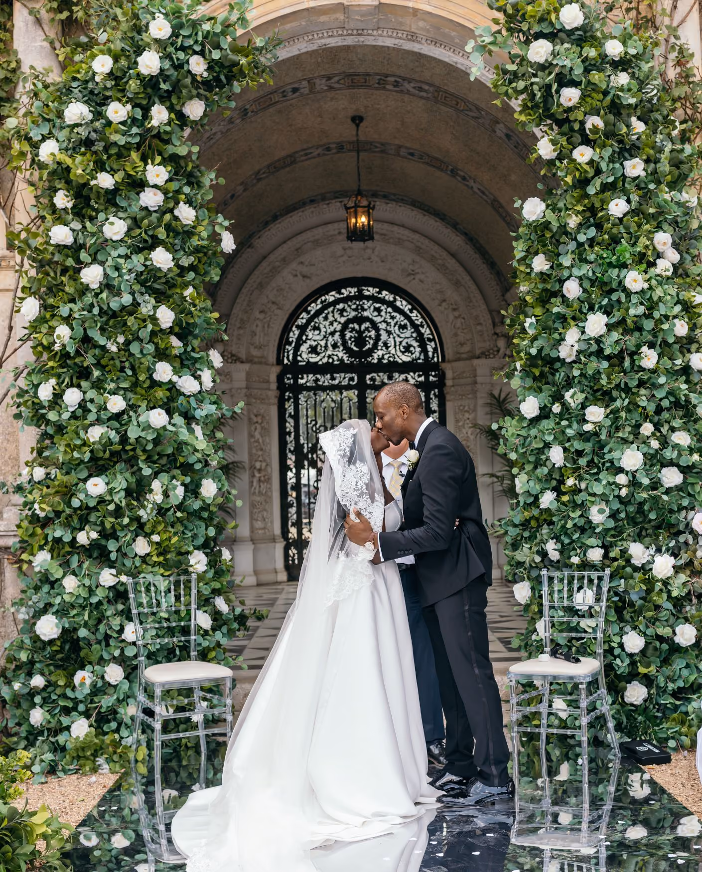 An Afro-Caribbean bride and groom sharing a kiss beneath a floral archway during their outdoor ceremony at North Mymms Park.