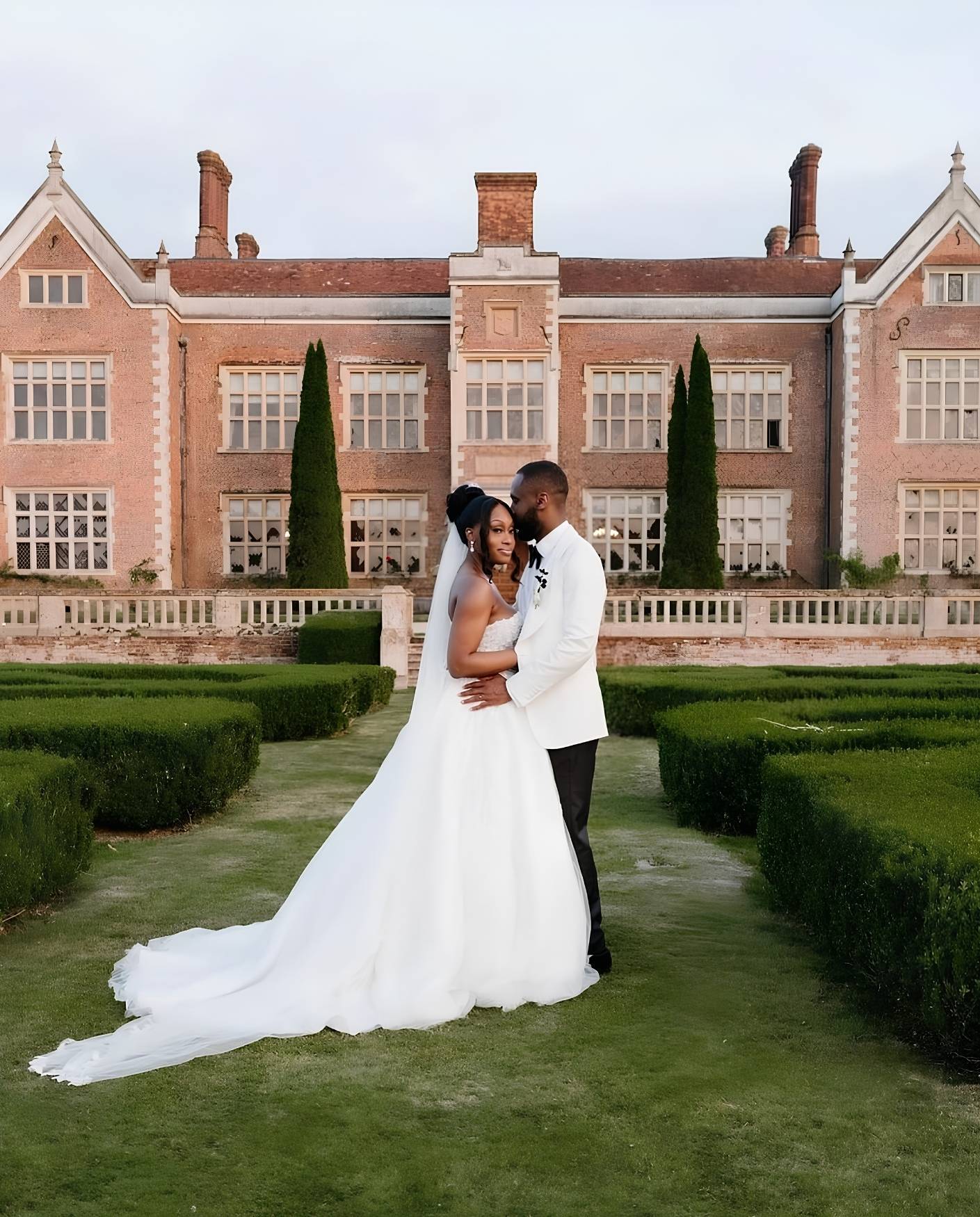 An African bride and groom embrace in the manicured gardens of North Mymms Park, with the historic red brick manor house forming the background.