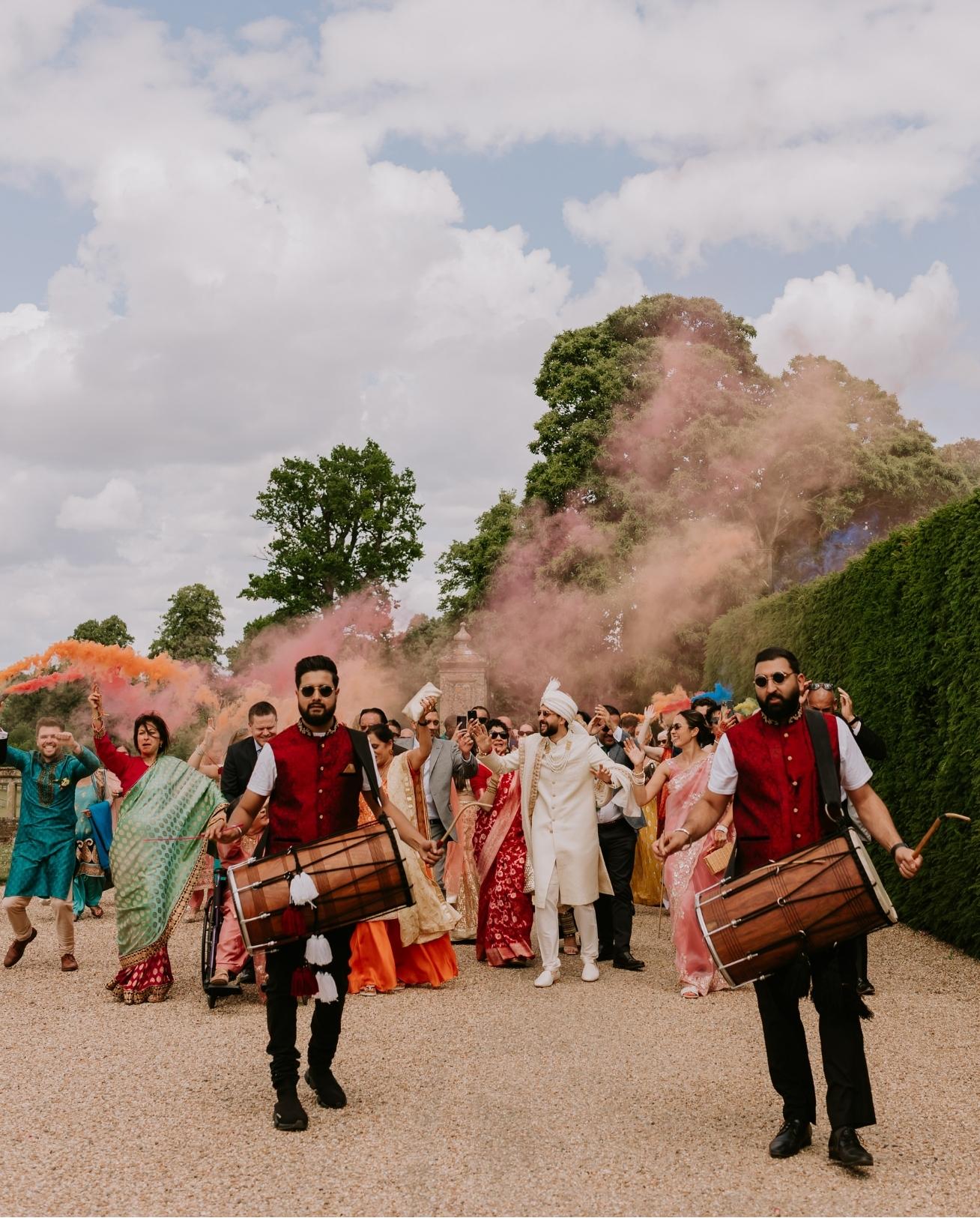 Dhol players lead a lively Baraat procession with colourful smoke flares outdoors at North Mymms Park.