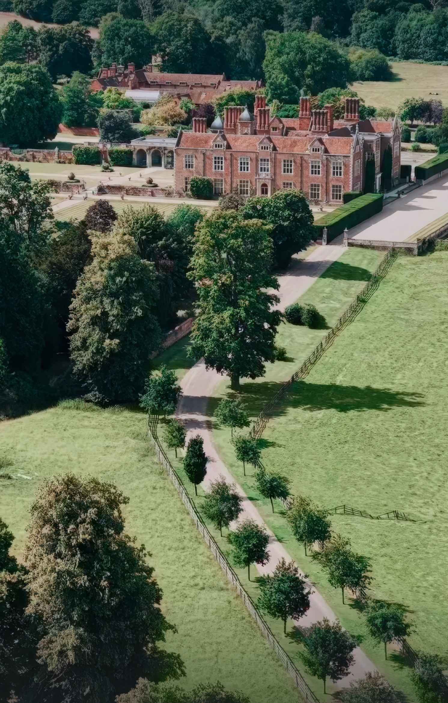An elevated aerial view showcasing the sprawling country estate of North Mymms Park. A long, winding private driveway lined with mature trees leads through green parkland towards the grand historic red-brick mansion and its surrounding gardens.