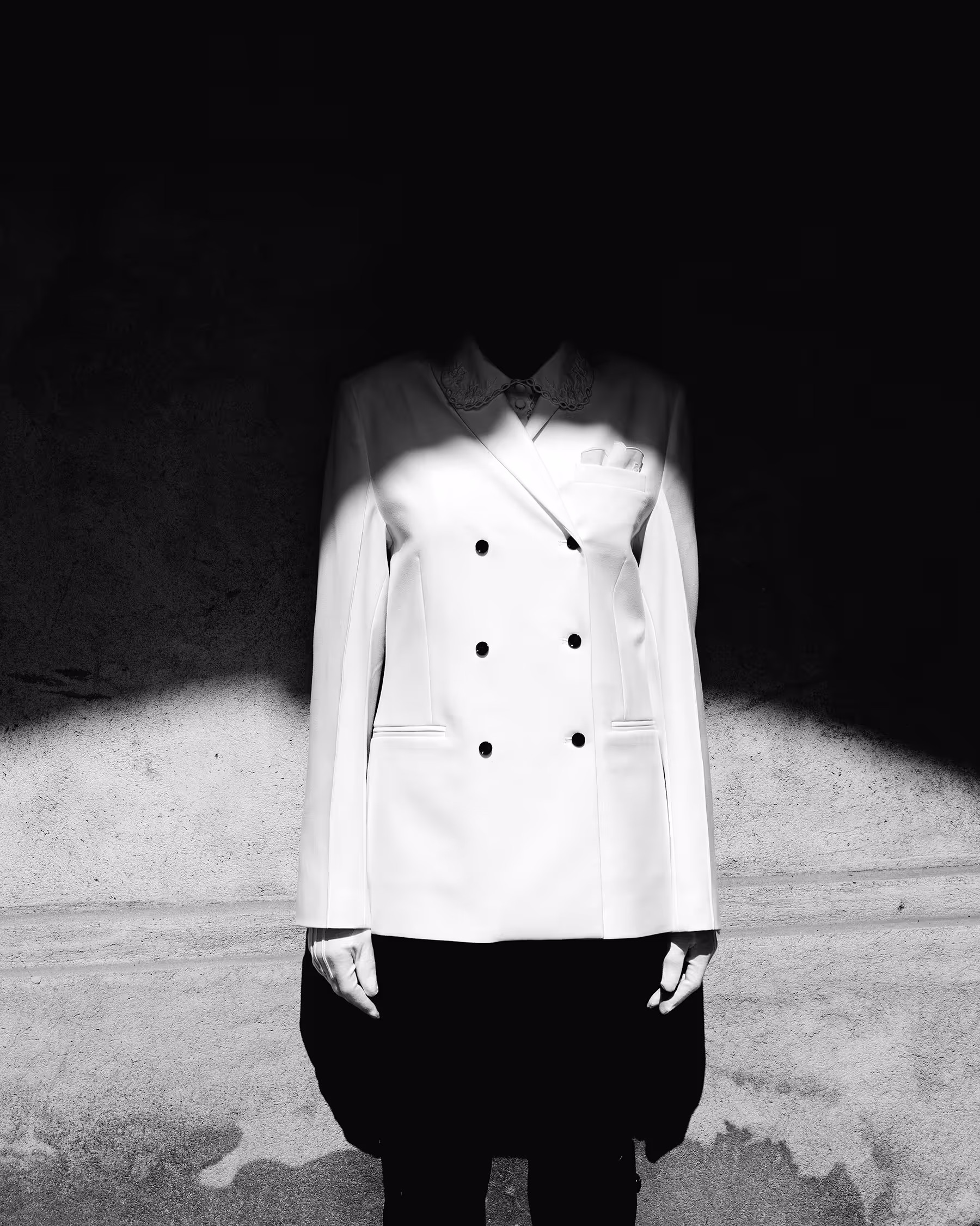 Artistic black and white runway photo from Copenhagen Fashion Week August 2025. A model wears a white double-breasted blazer with their face obscured by dramatic shadow.
