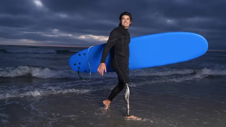 Smiling man with a prosthetic leg carrying a blue surfboard walking on the beach at dusk with waves in the background.