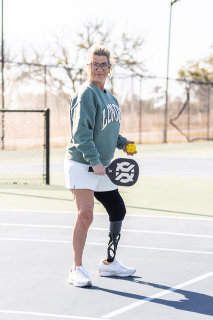 Woman with a prosthetic leg holding a pickleball paddle and ball on an outdoor court.
