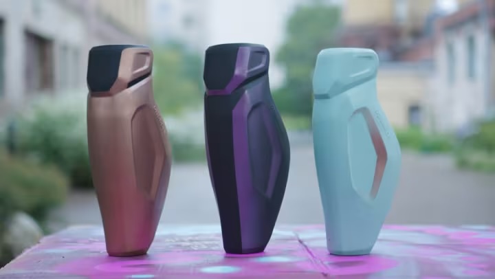 Three modern, ergonomic water bottles in bronze, black-purple, and light blue colors placed on a pink surface outdoors.