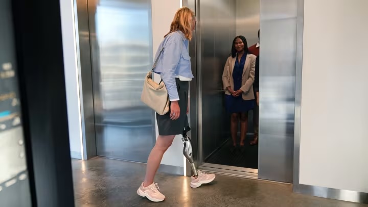A woman with a prosthetic leg is walking towards an open elevator door where two people stand inside.