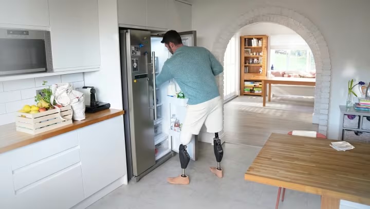 Man with bilateral prosthetic legs reaching into an open refrigerator in a modern white kitchen.