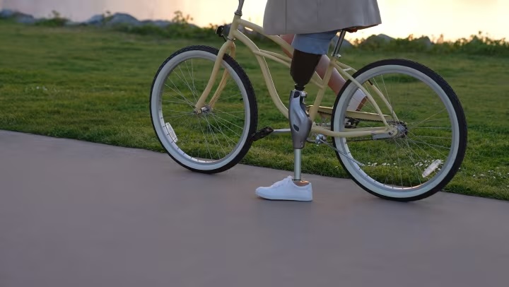 Person with a prosthetic leg standing next to a beige bicycle on a paved path with grass in the background.