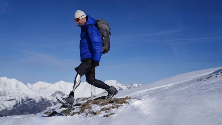 Person with a prosthetic leg wearing a blue jacket and gray backpack hiking on a snowy mountain slope under a clear blue sky.
