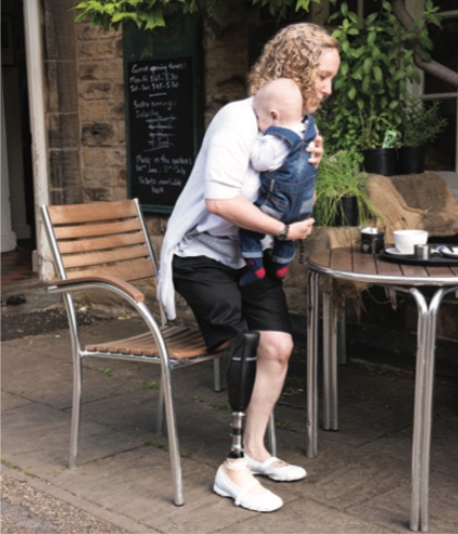 Person with a prosthetic leg sitting on a chair holding a baby, outdoors at a café table.