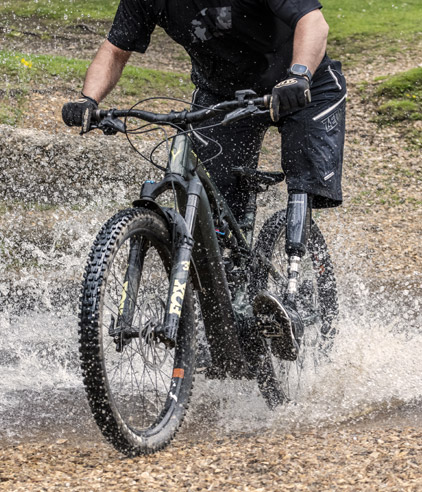 Person with a prosthetic leg riding a mountain bike through splashing water on a dirt trail.