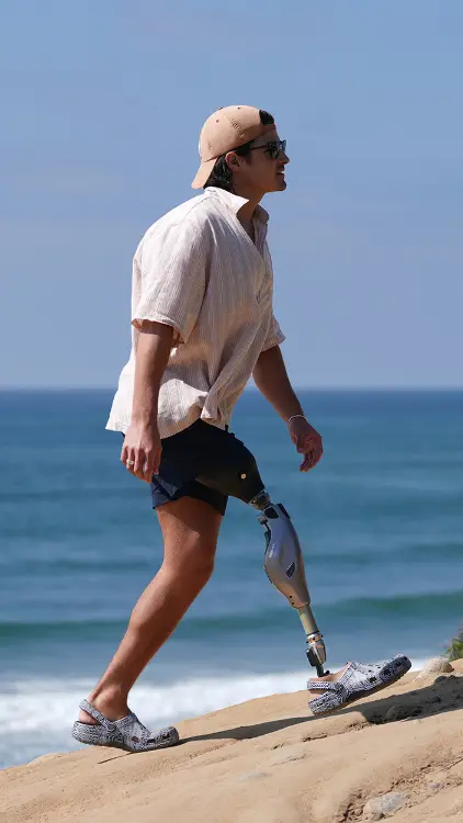 Man with prosthetic leg walking on a sandy beach uphill wearing a cap and sunglasses.