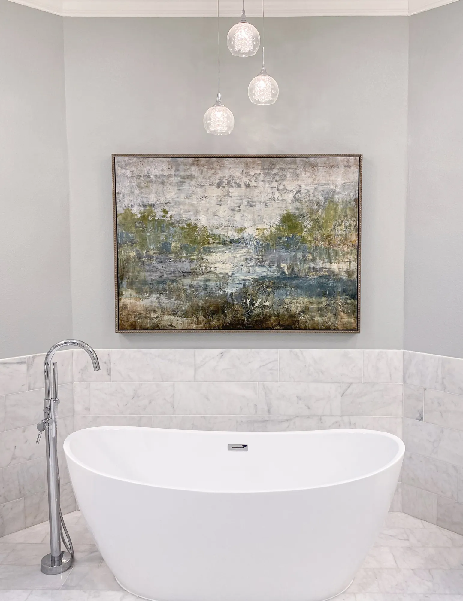 Modern bathroom with white freestanding bathtub, chrome floor-mounted faucet, abstract landscape painting, and three pendant lights.
