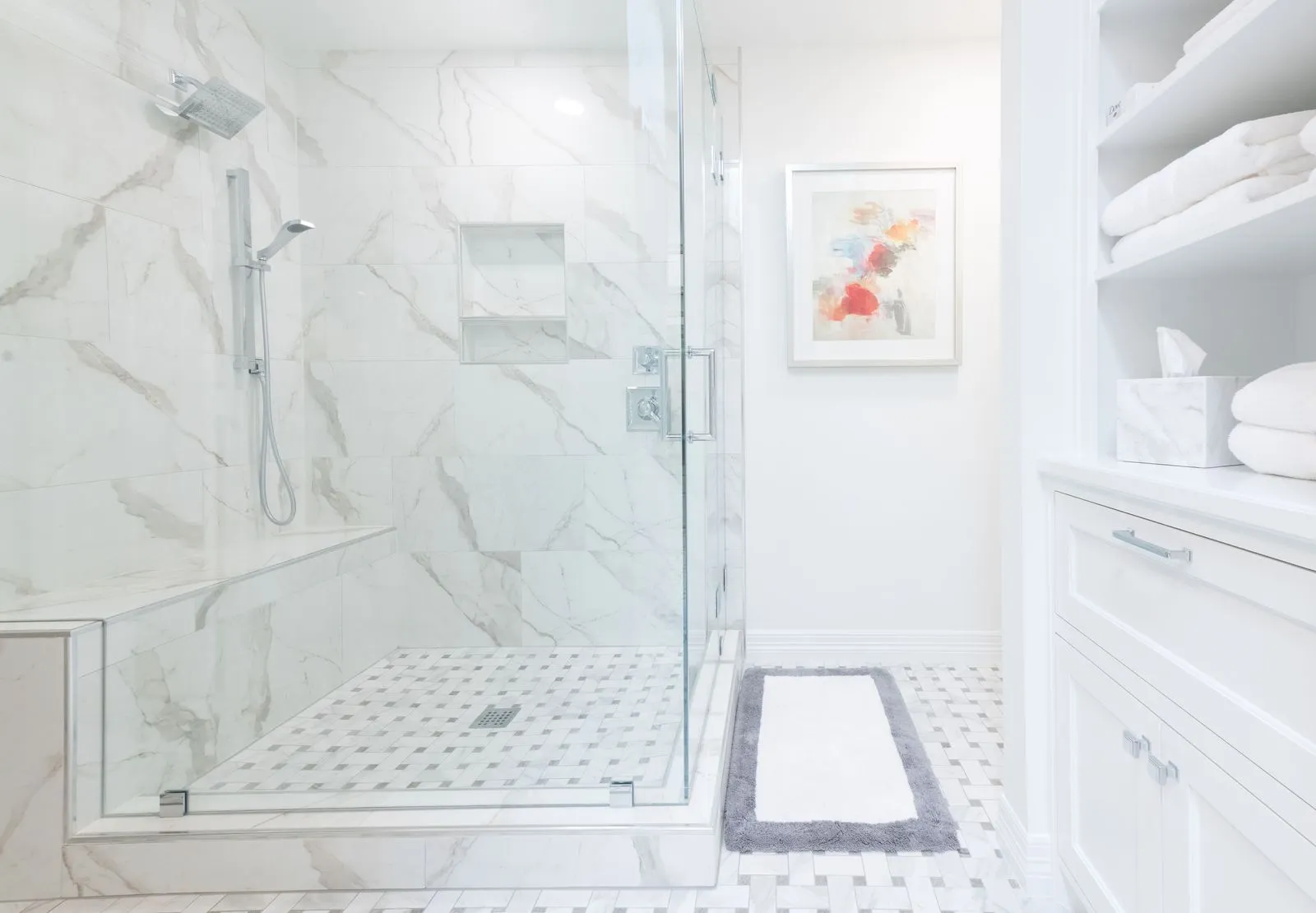 Bright white bathroom featuring a glass-enclosed marble tiled shower with built-in bench and adjacent shelves with folded towels.
