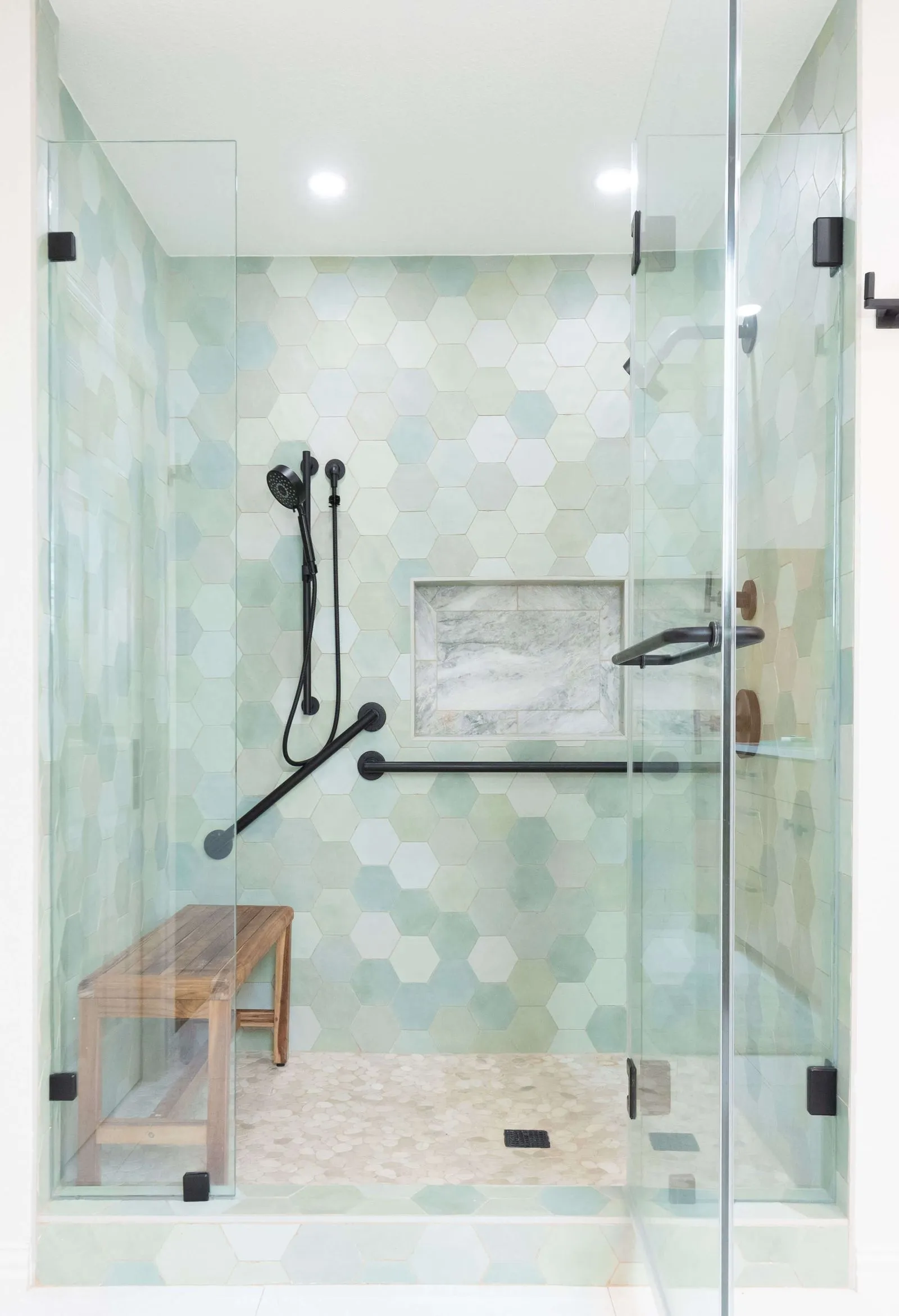 Modern walk-in shower with pastel hexagonal wall tiles, black fixtures, a wooden bench, and glass doors.