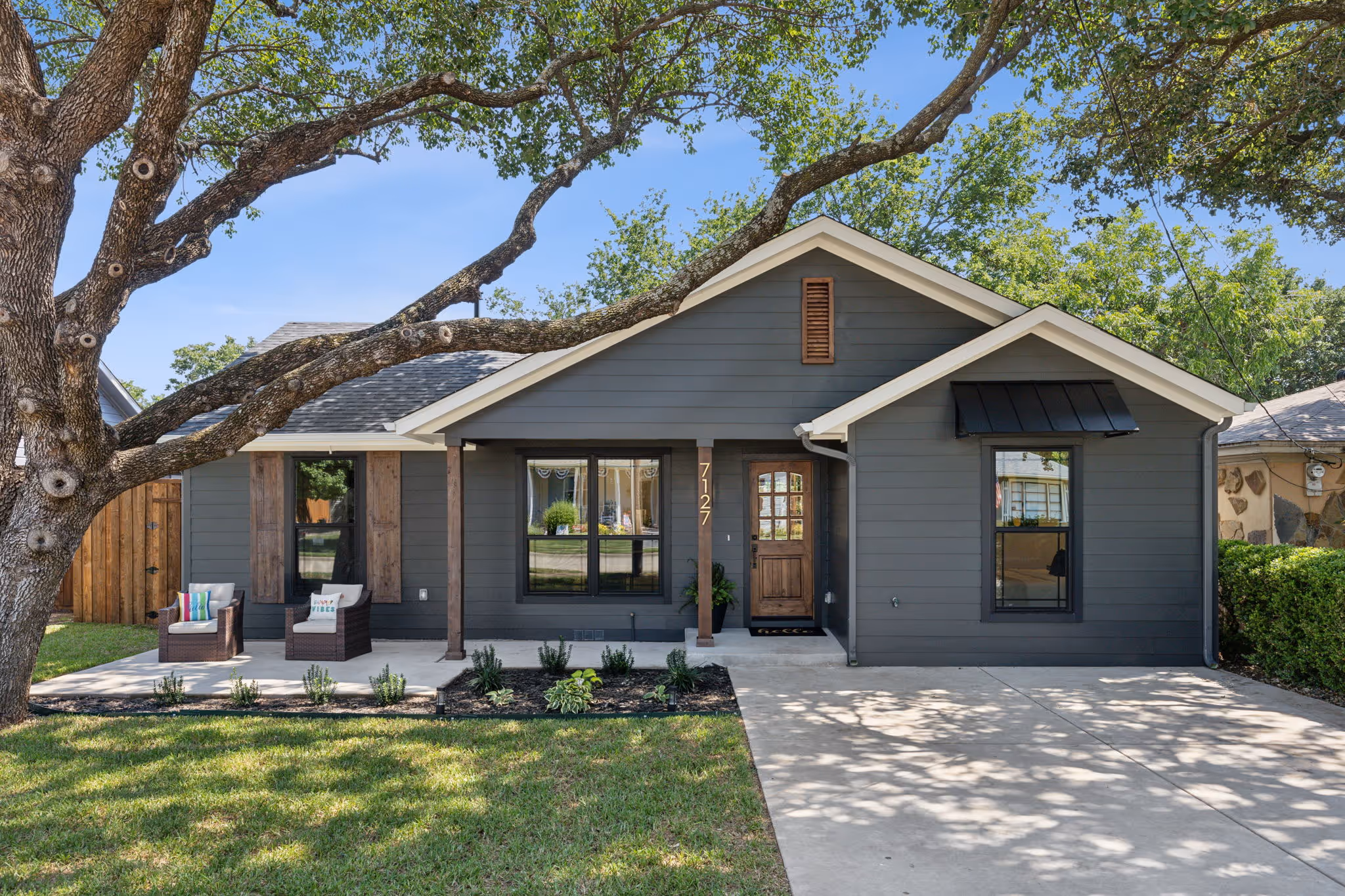 Gray single-story house with wooden front door, large tree with sprawling branches in front, and a small porch with two chairs.