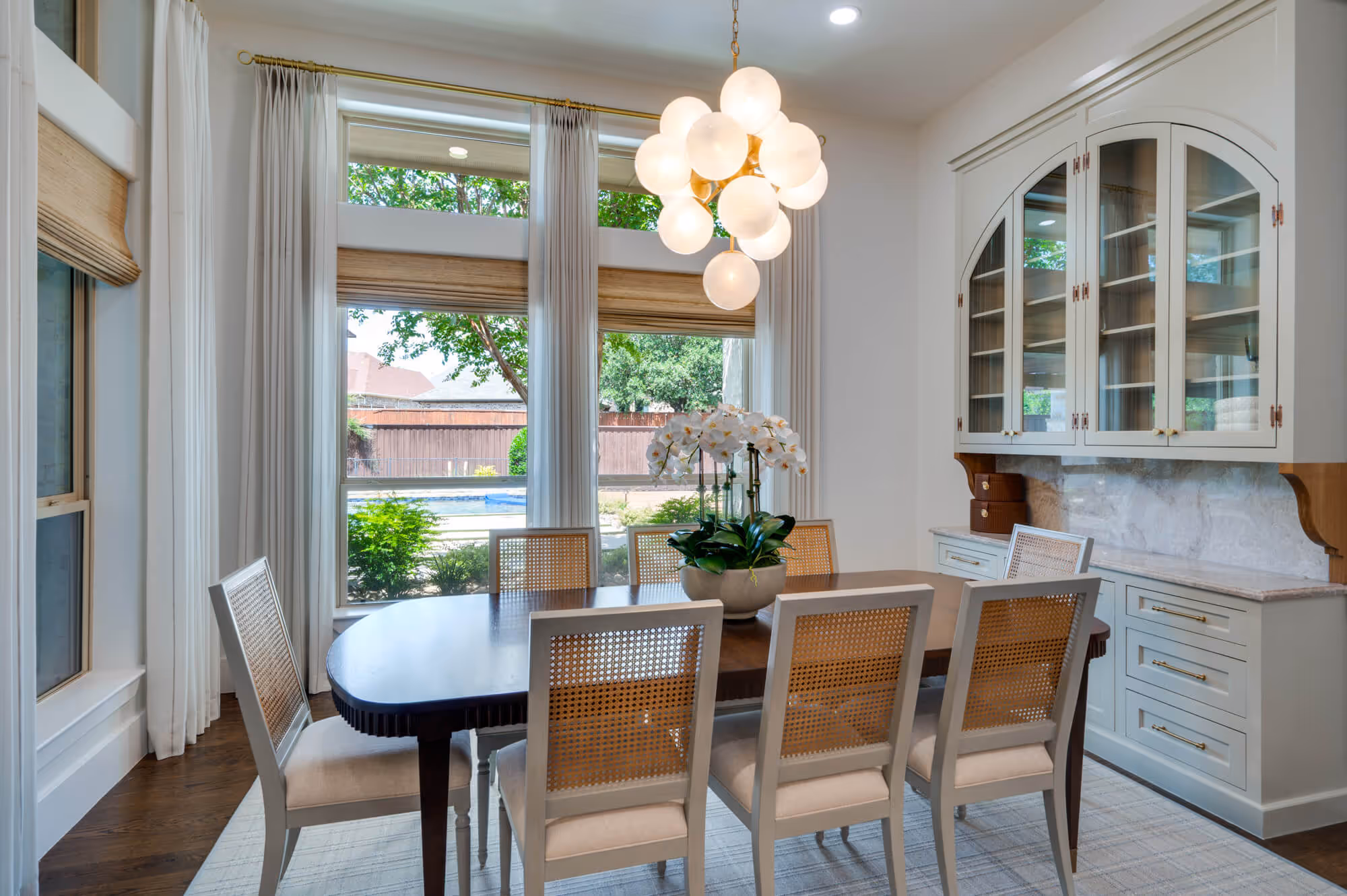 Modern dining room with a dark wood table surrounded by eight light-colored chairs with cane backs, a white floral centerpiece, large windows with curtains, and a white cabinet with glass doors.