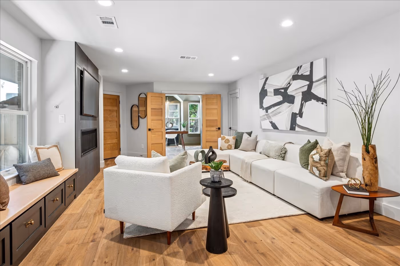 Bright modern living room with white sofa, armchair, wooden floor, abstract black and white painting, and a small round table with a plant.