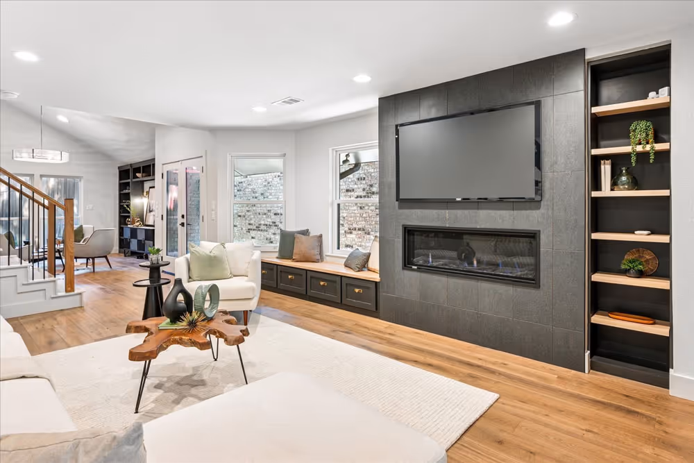 Modern living room with a mounted flat-screen TV above a sleek fireplace, wood flooring, white furniture, and built-in shelves with decorative items.