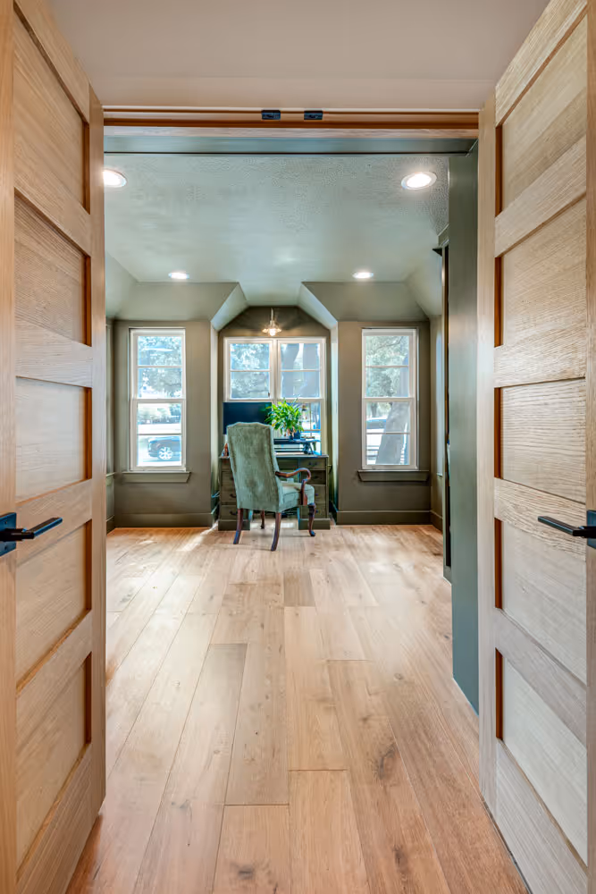 Interior view through open wooden double doors into a modern room with light wood flooring, three windows and a single green upholstered chair facing a small desk with a plant on it.