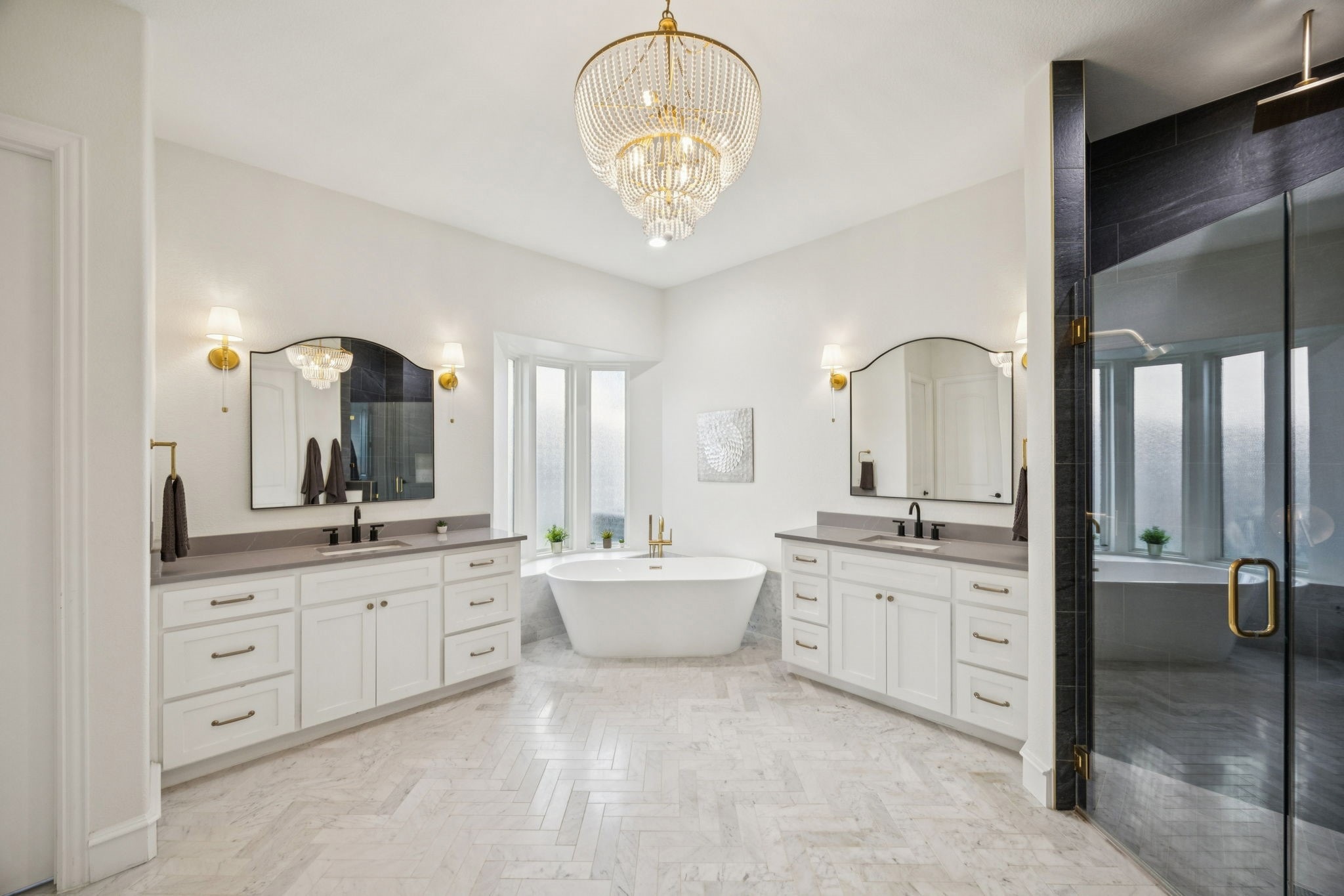 Luxury bathroom with white double vanities, freestanding tub, large window, chandelier, and glass shower.