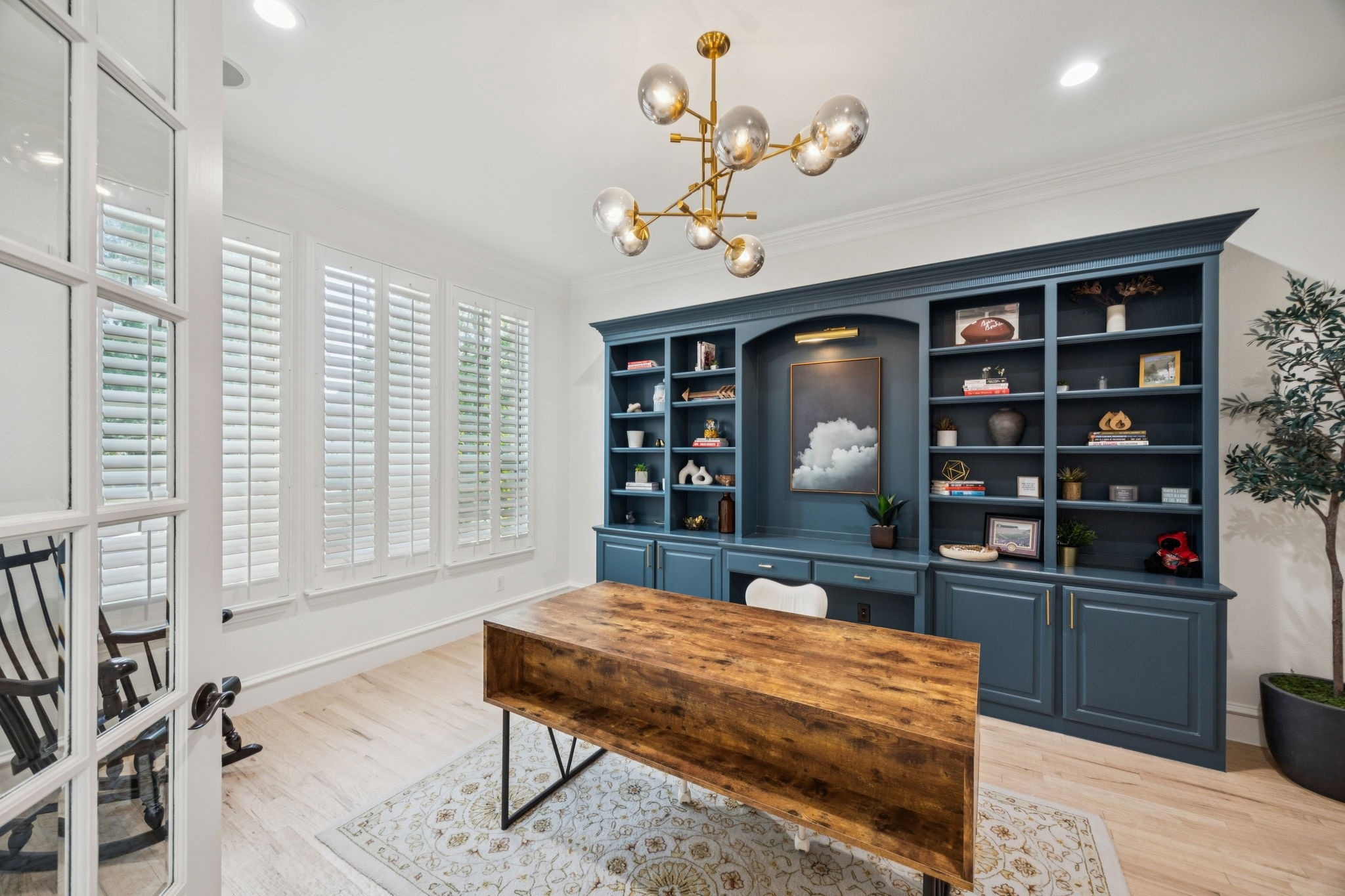 Home office with large wooden desk, blue built-in shelves, white walls, and modern chandelier.