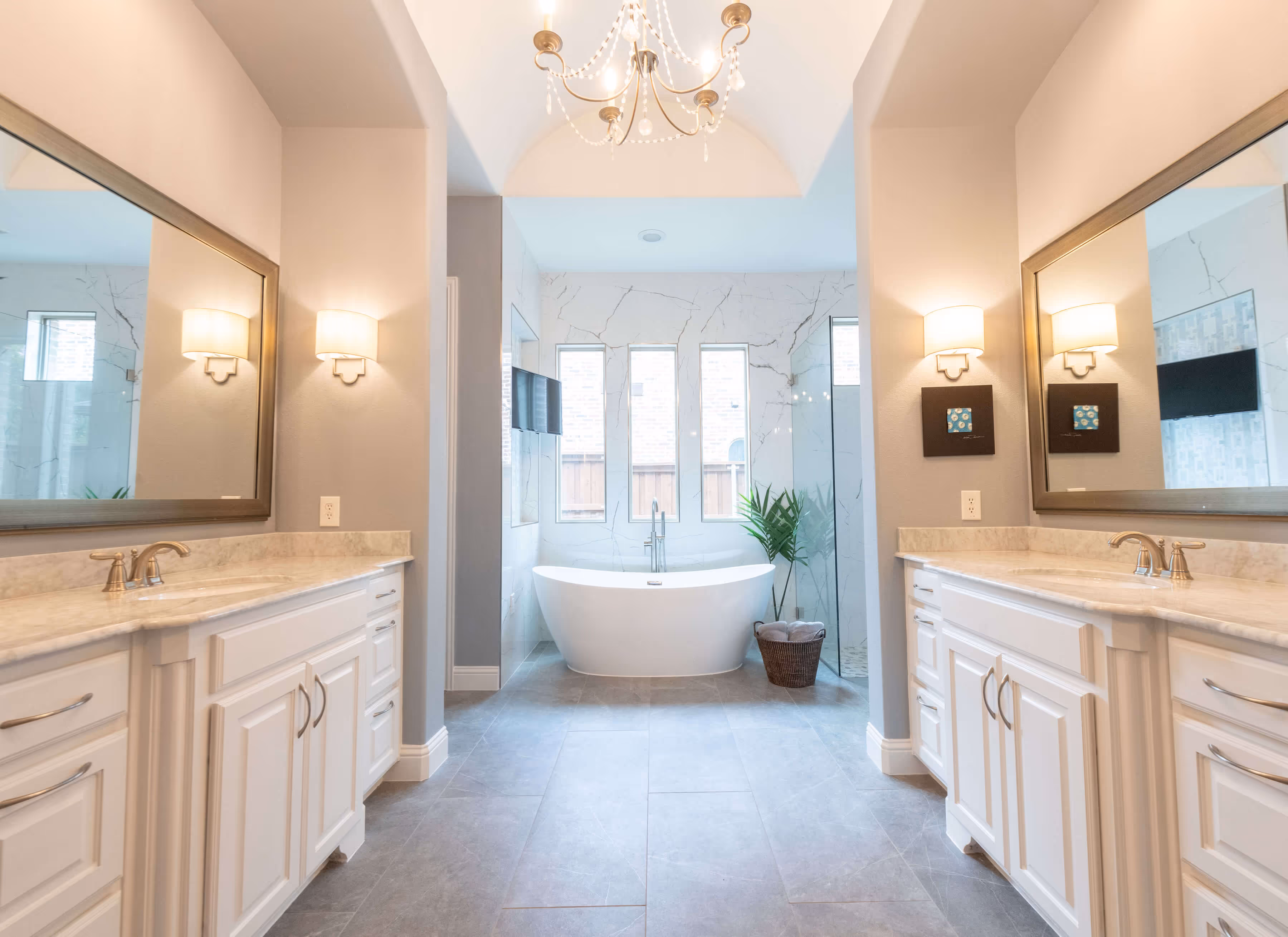 Luxury bathroom with white double vanities, freestanding tub, large window, chandelier, and glass shower.