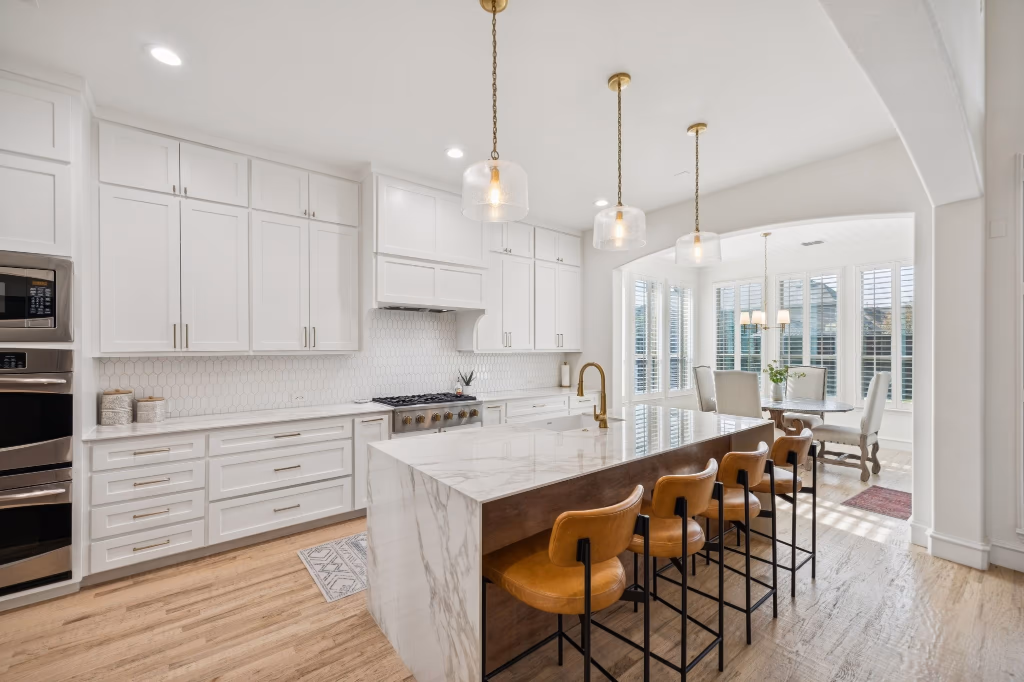 Modern kitchen with white cabinets, marble island, tan bar stools, and dining nook with large windows.