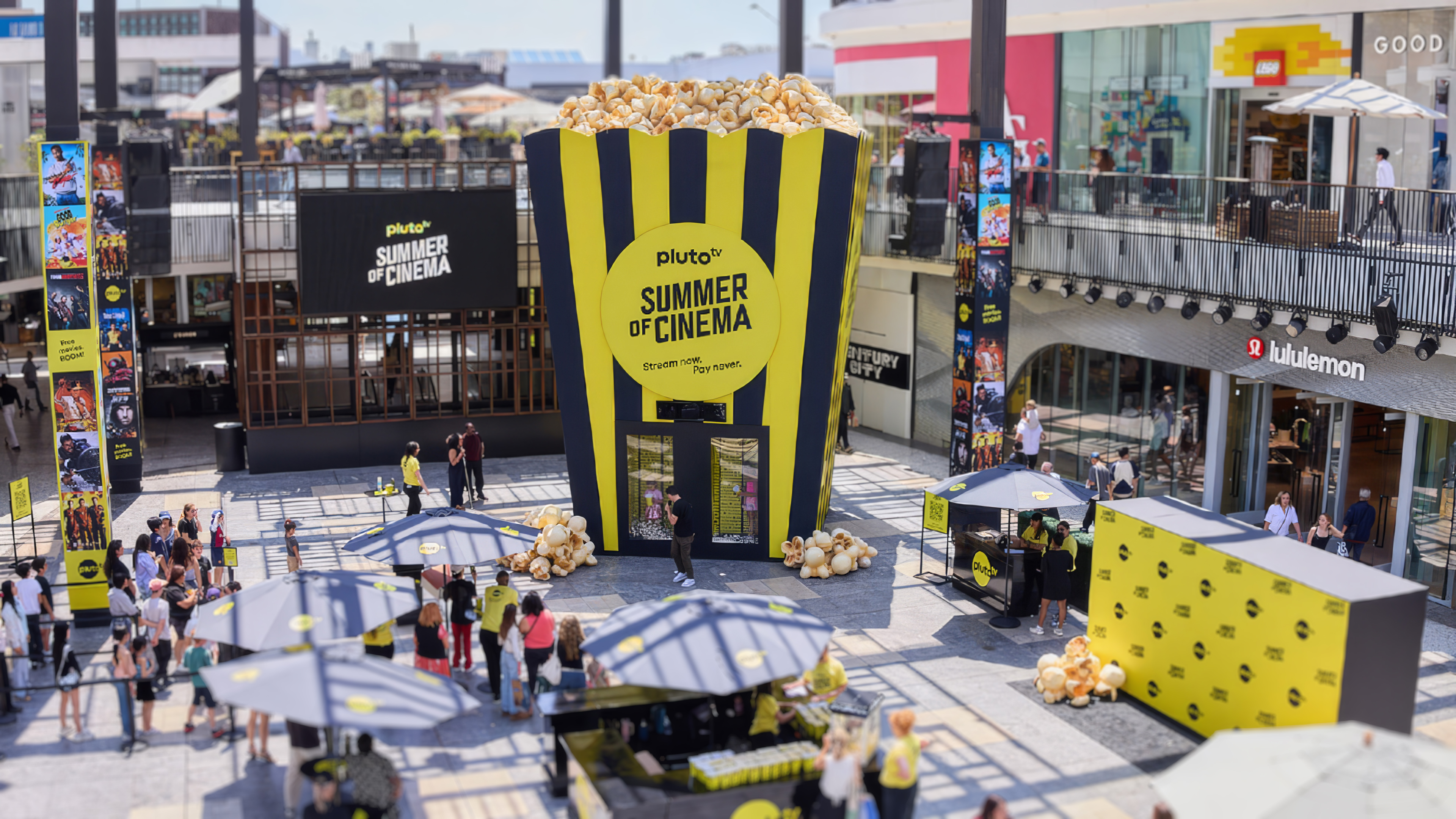 Outdoor event space with a giant popcorn bucket installation promoting Pluto TV Summer of Cinema, surrounded by people and branded tents.