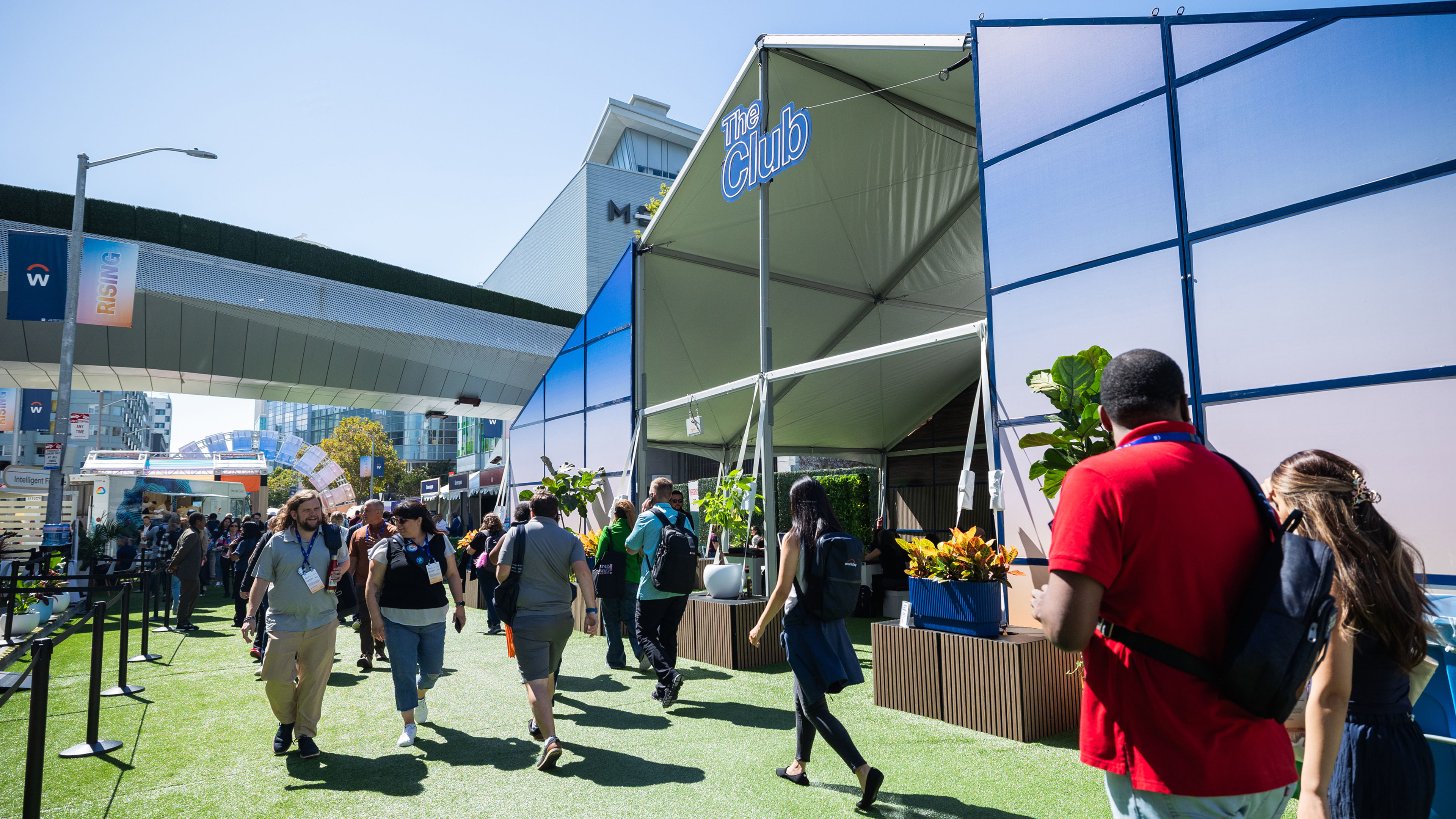 People walking along an outdoor green-carpeted event area with a tent labeled 'The Club' and large blue panels.