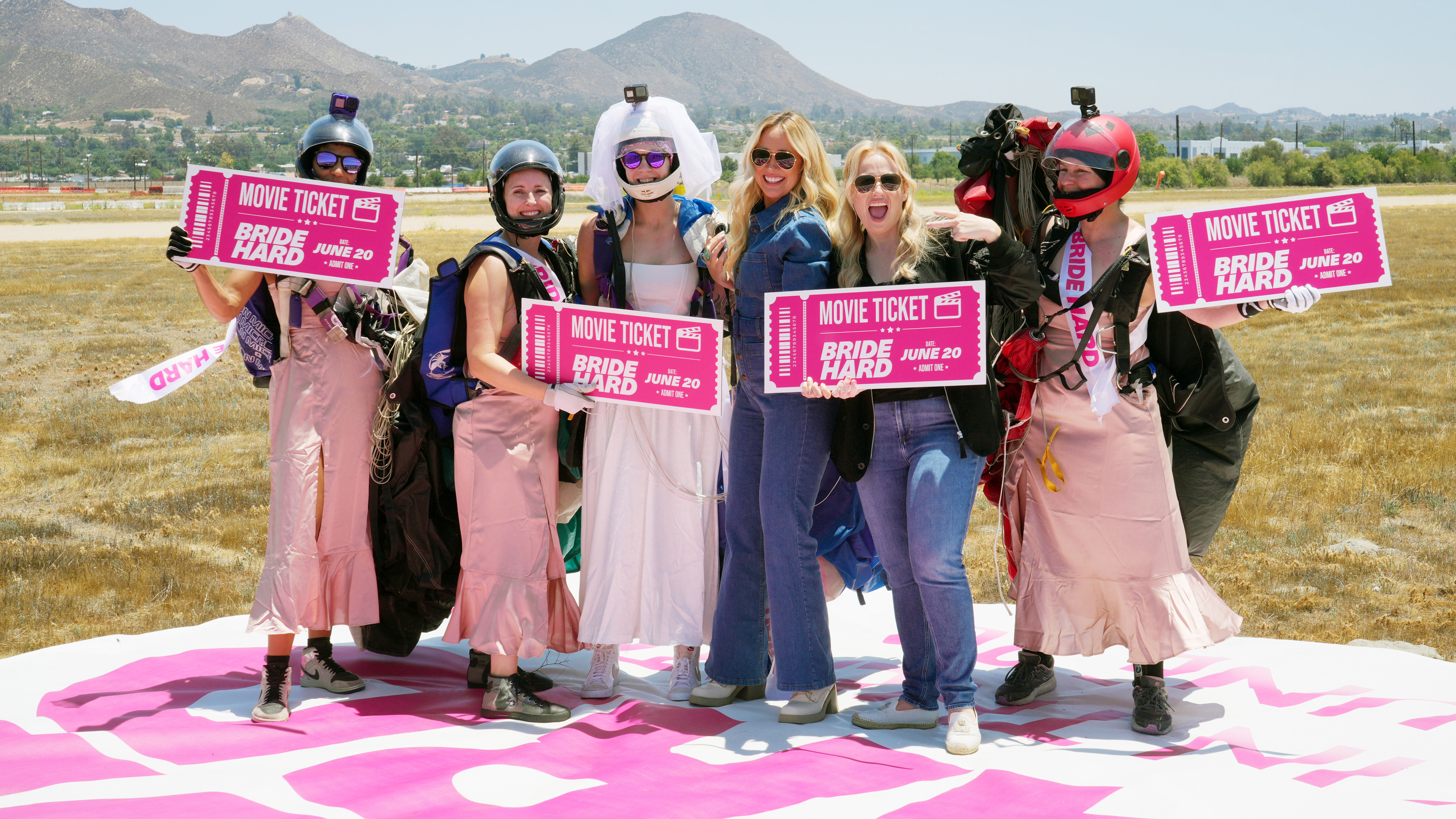 Group of women outdoors holding pink Bride Hard movie ticket signs, some wearing helmets and parachutes with mountainous background.