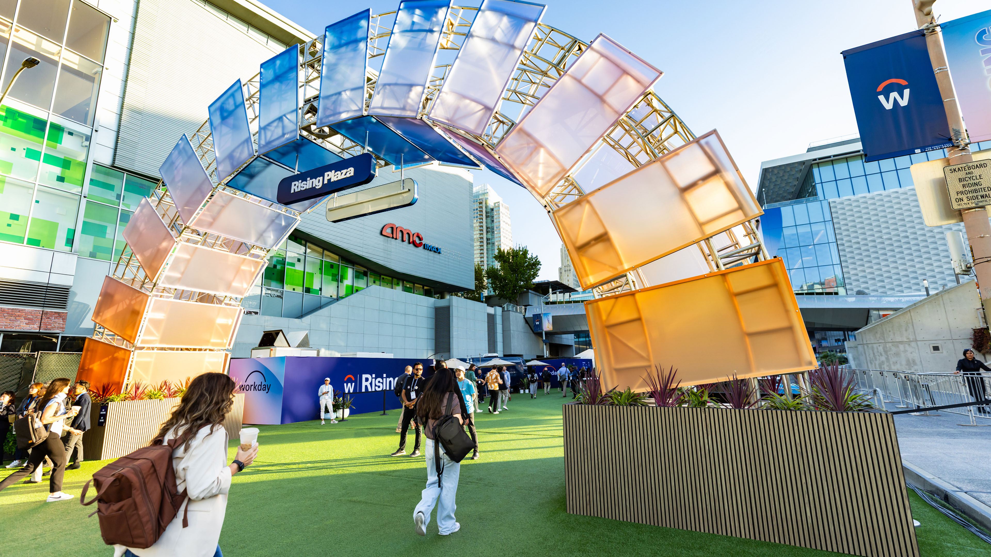 Outdoor plaza with people walking under a colorful arch installation labeled 'Rising Plaza' near a building with AMC IMAX signage.