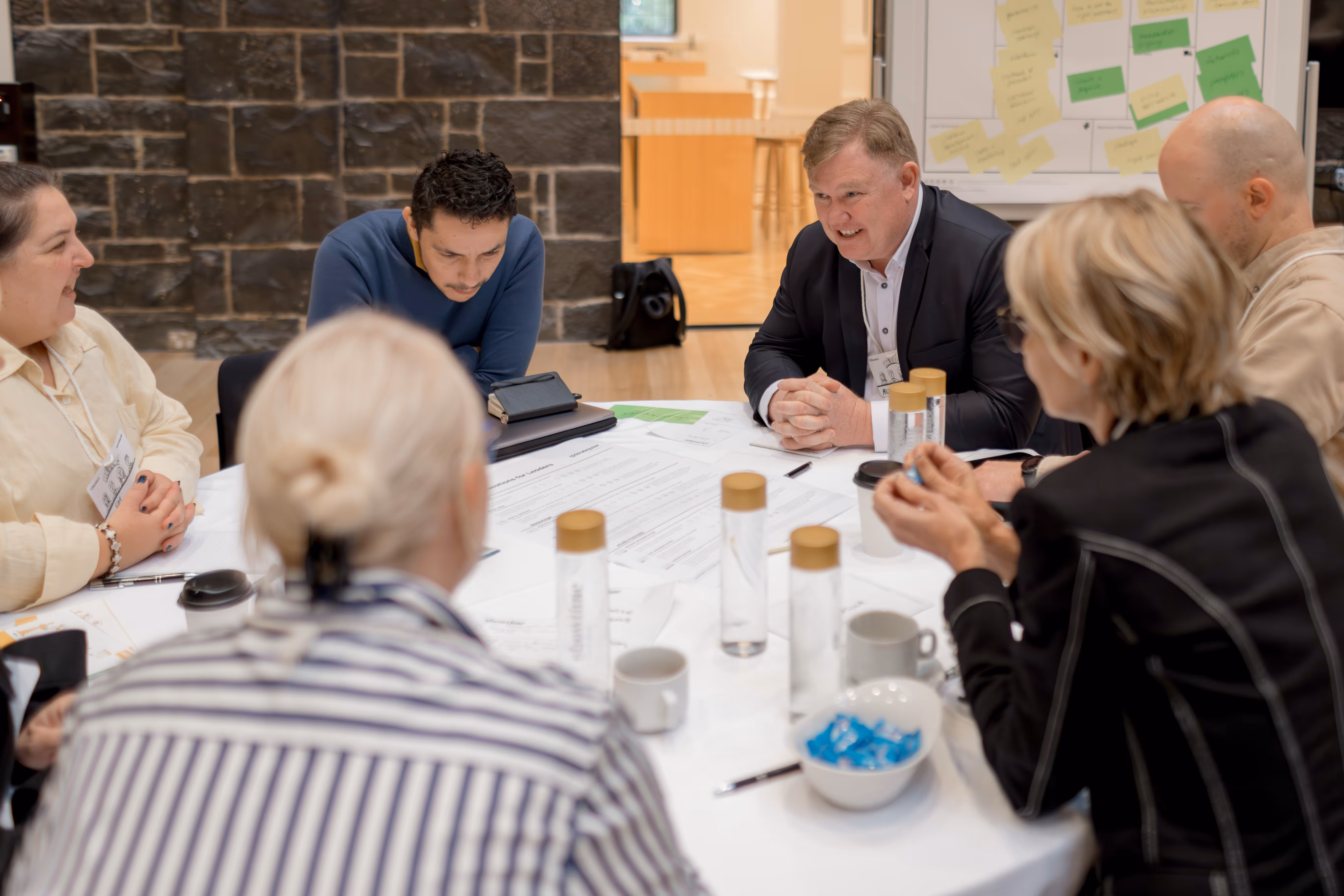 Six people engaged in a business meeting around a round table covered with documents, water bottles, and coffee cups.
