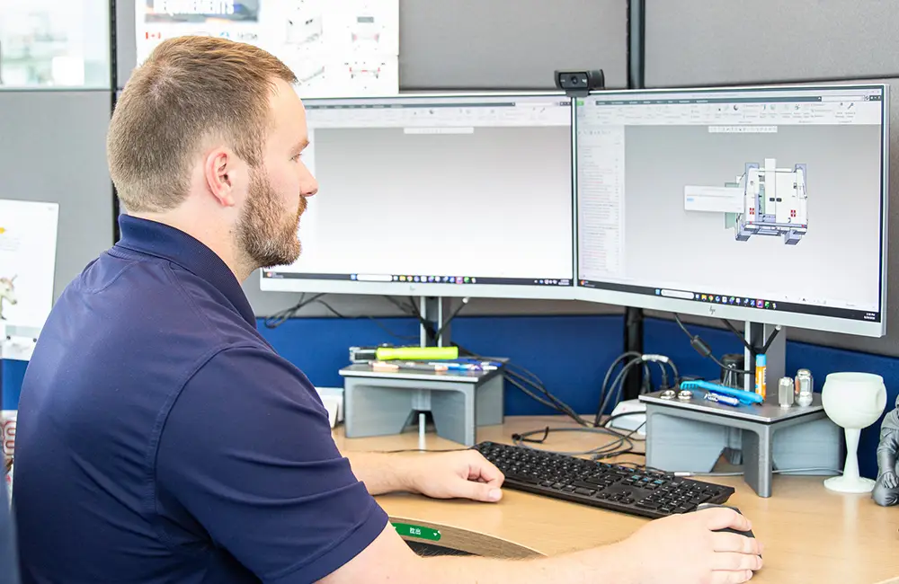 A man at a dual-monitor workstation reviews a 3D CAD model of a truck body design.