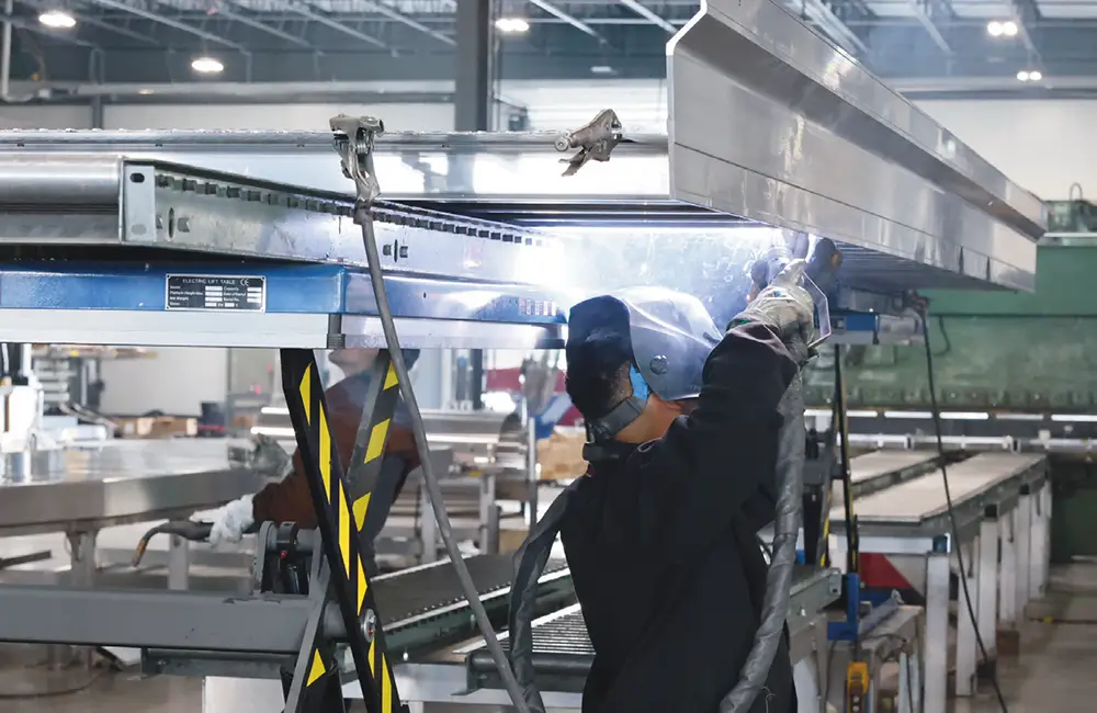 A worker in a welding helmet and protective gear welds an aluminum trailer frame component on a production line.