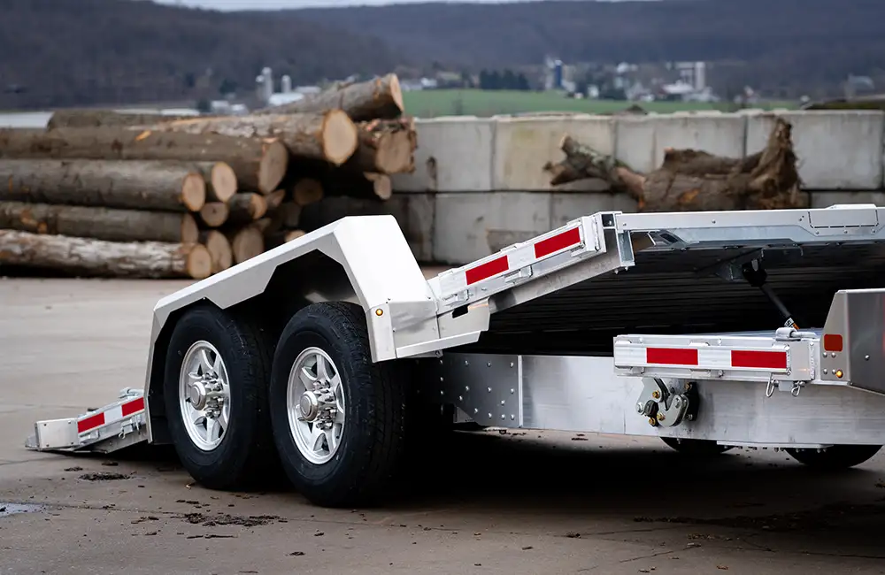 An Eby GT14K tilt-bed aluminum equipment trailer with its deck angled down for loading, parked near stacked logs.