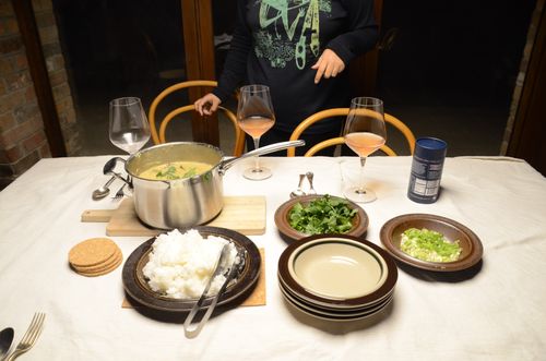 Close up of a dining table with a light canvas table cloth. There's a big silver bowl filled with thai green curry, a bowl of rice, some greens and wine glasses.