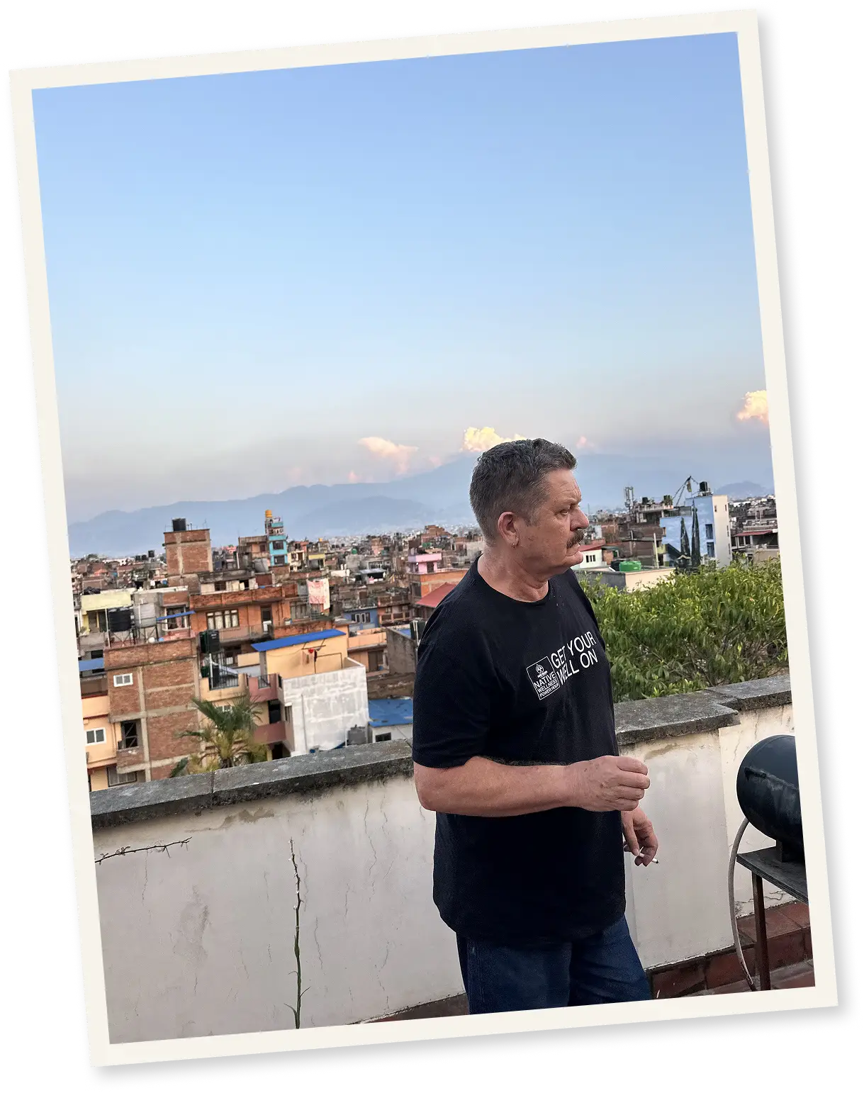 Man in black shirt standing on rooftop with cityscape and mountains in background at sunset.