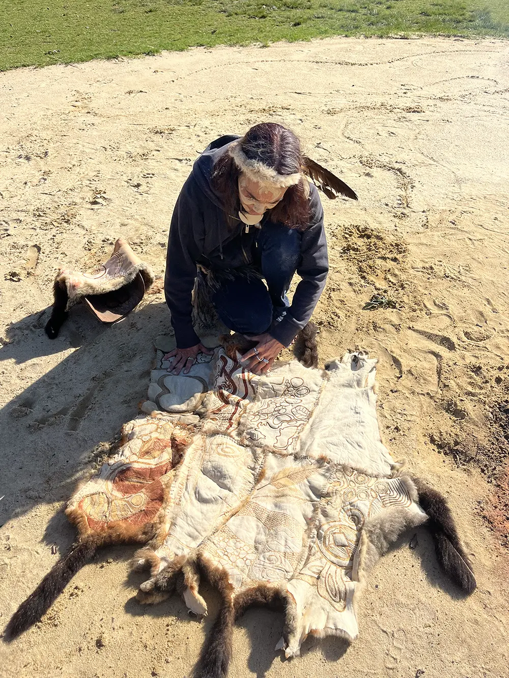 Person kneeling outdoors on dirt, touching animal hides with intricate patterns, wearing a feathered headband.